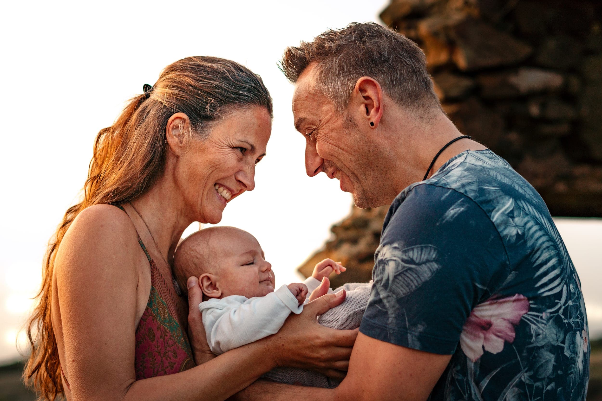 Parents gazing at each other while cradling their smiling newborn at sunset