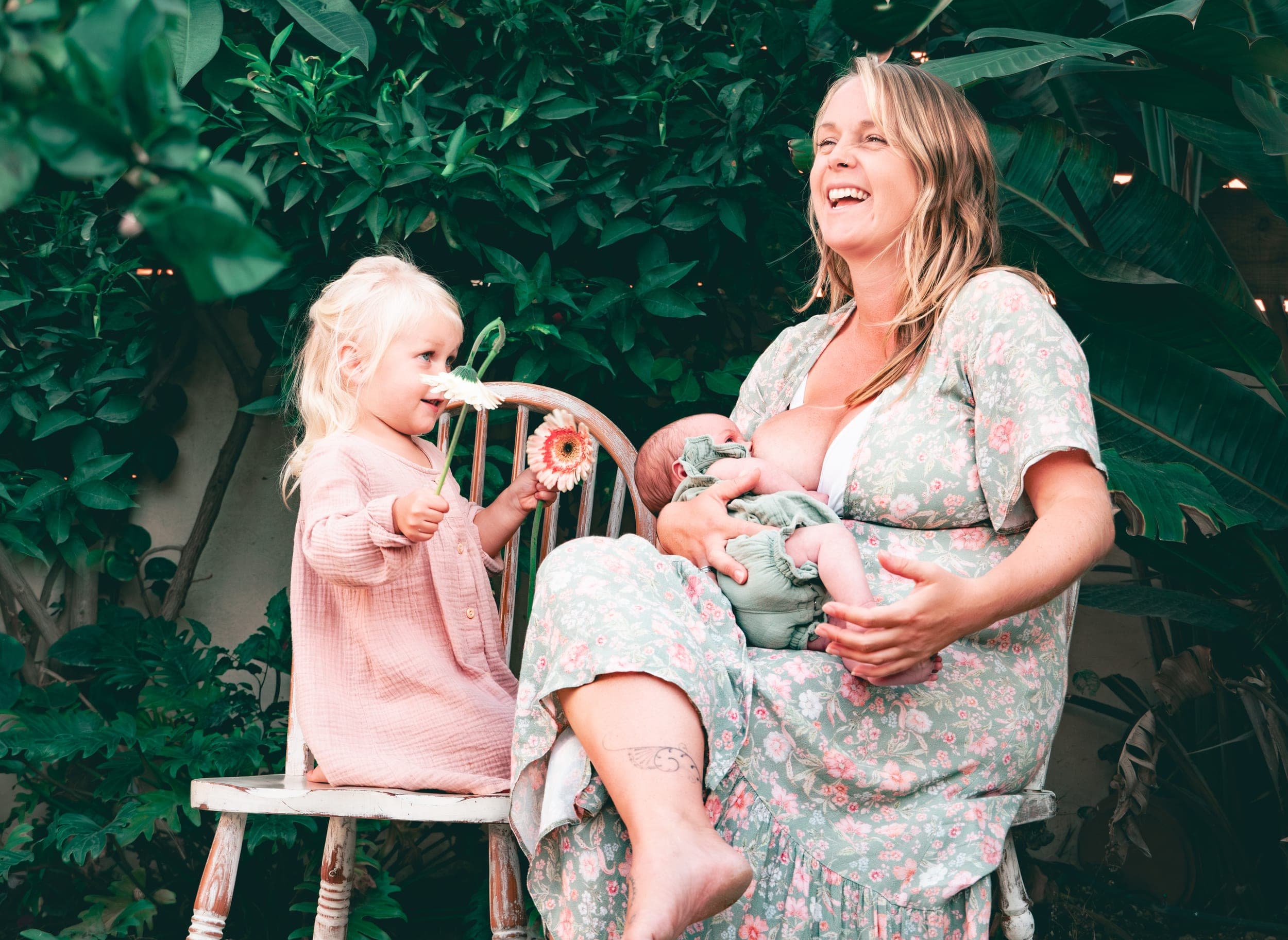 Laughing mother breastfeeding newborn while toddler holds flowers on a garden chair
