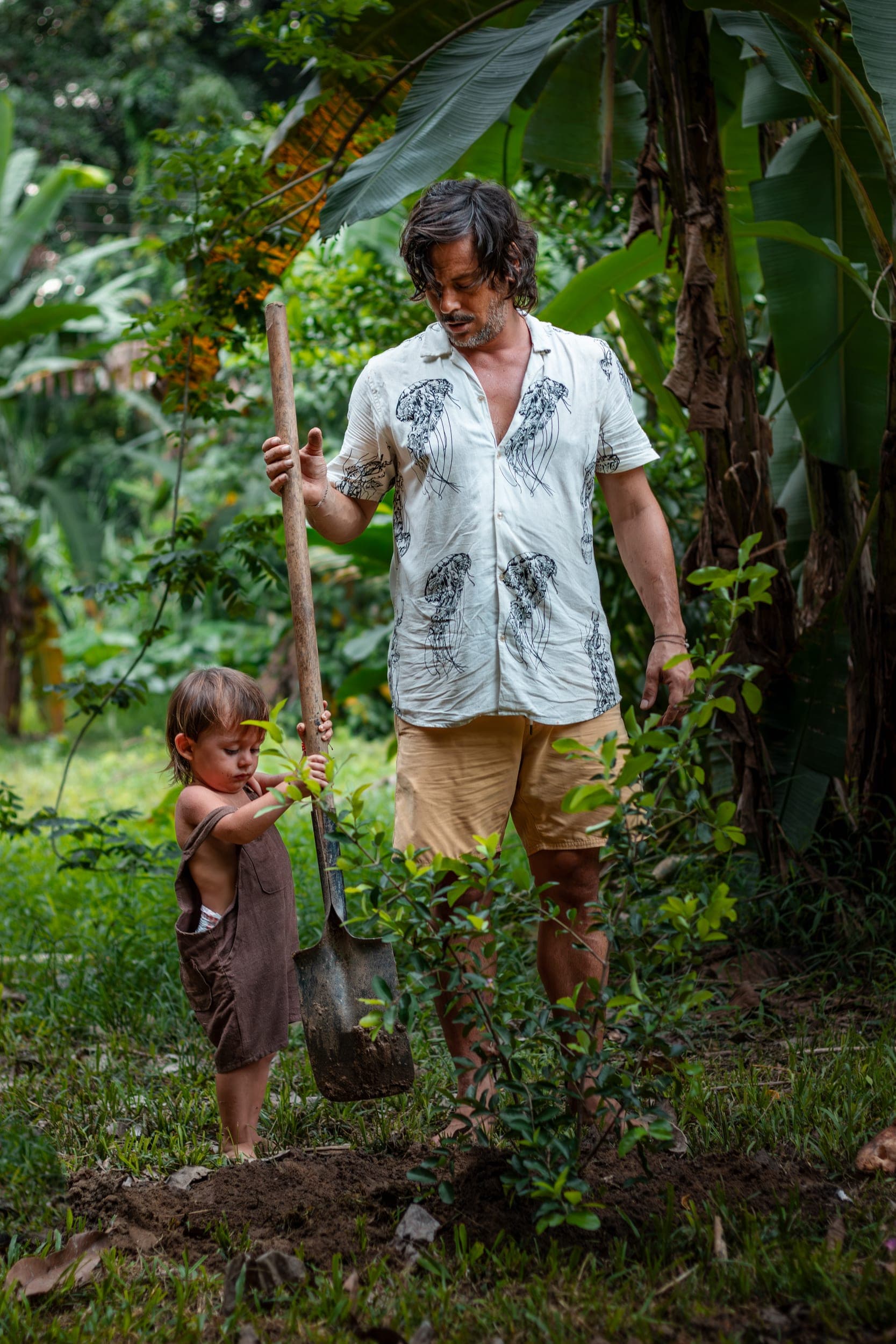 Father and barefoot toddler holding a shovel while planting in a jungle garden