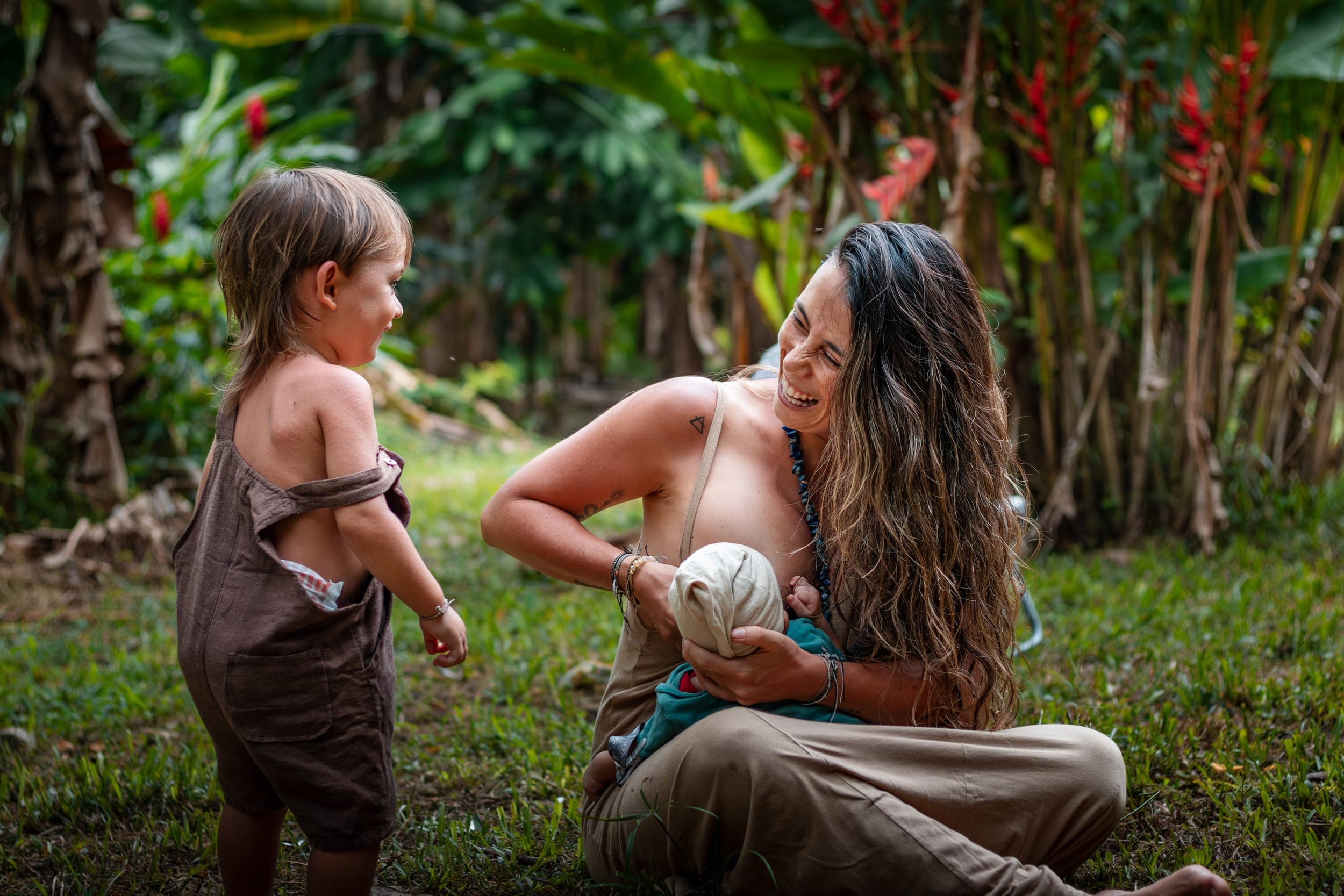 Laughing mother sitting on grass nursing baby as toddler stands beside her in garden