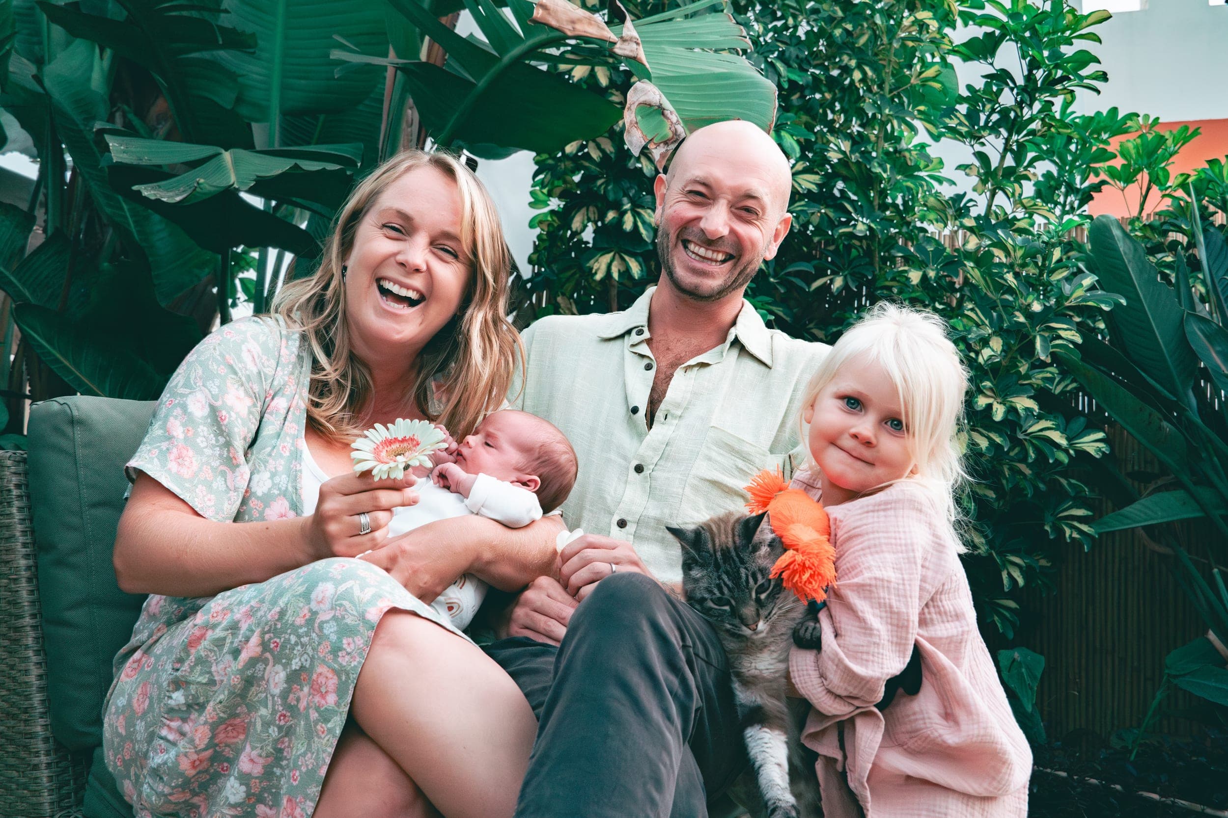 Laughing parents with newborn, young daughter and cat in lush tropical garden