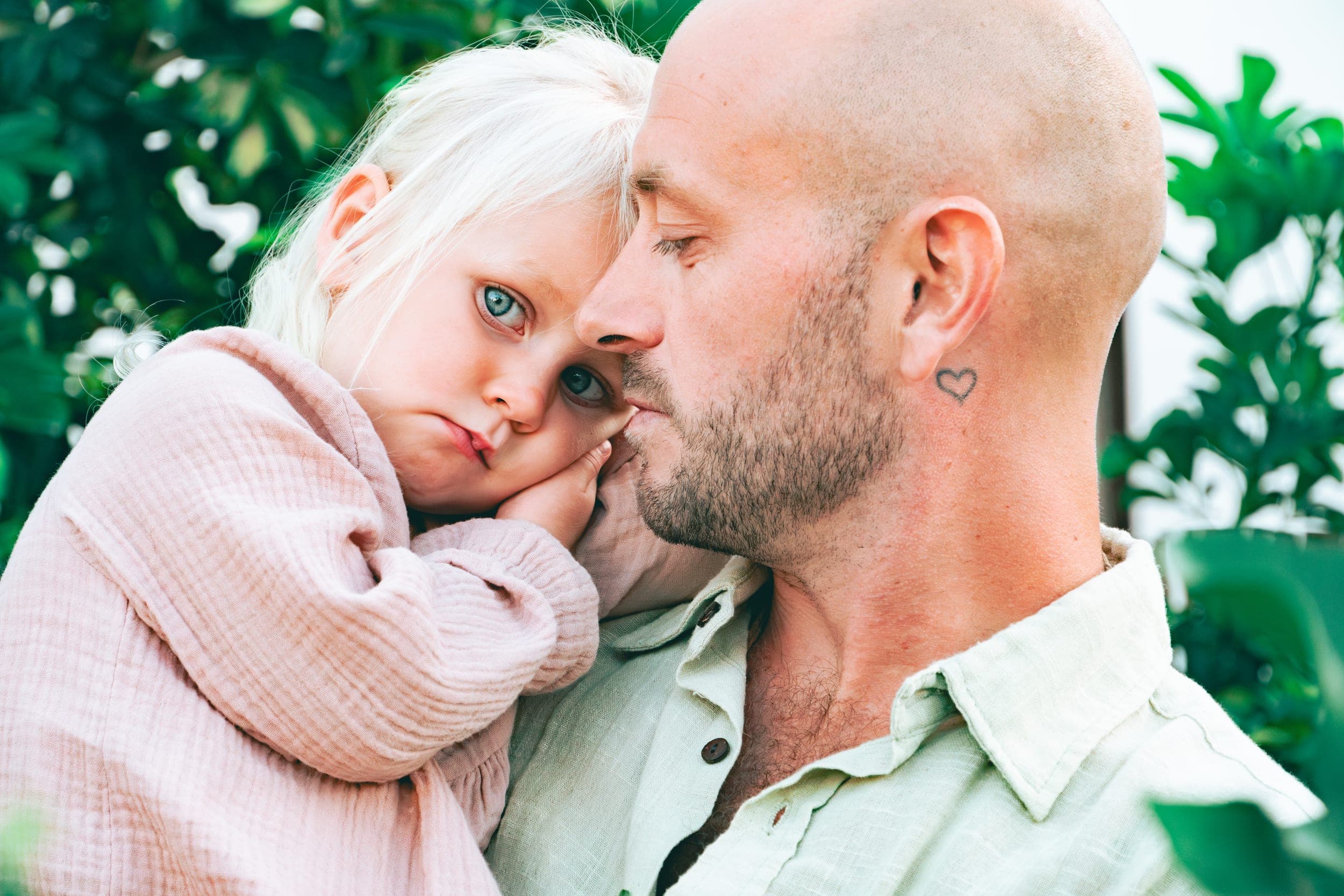 Bald father tenderly nuzzling his blonde daughter cheek to cheek among greenery