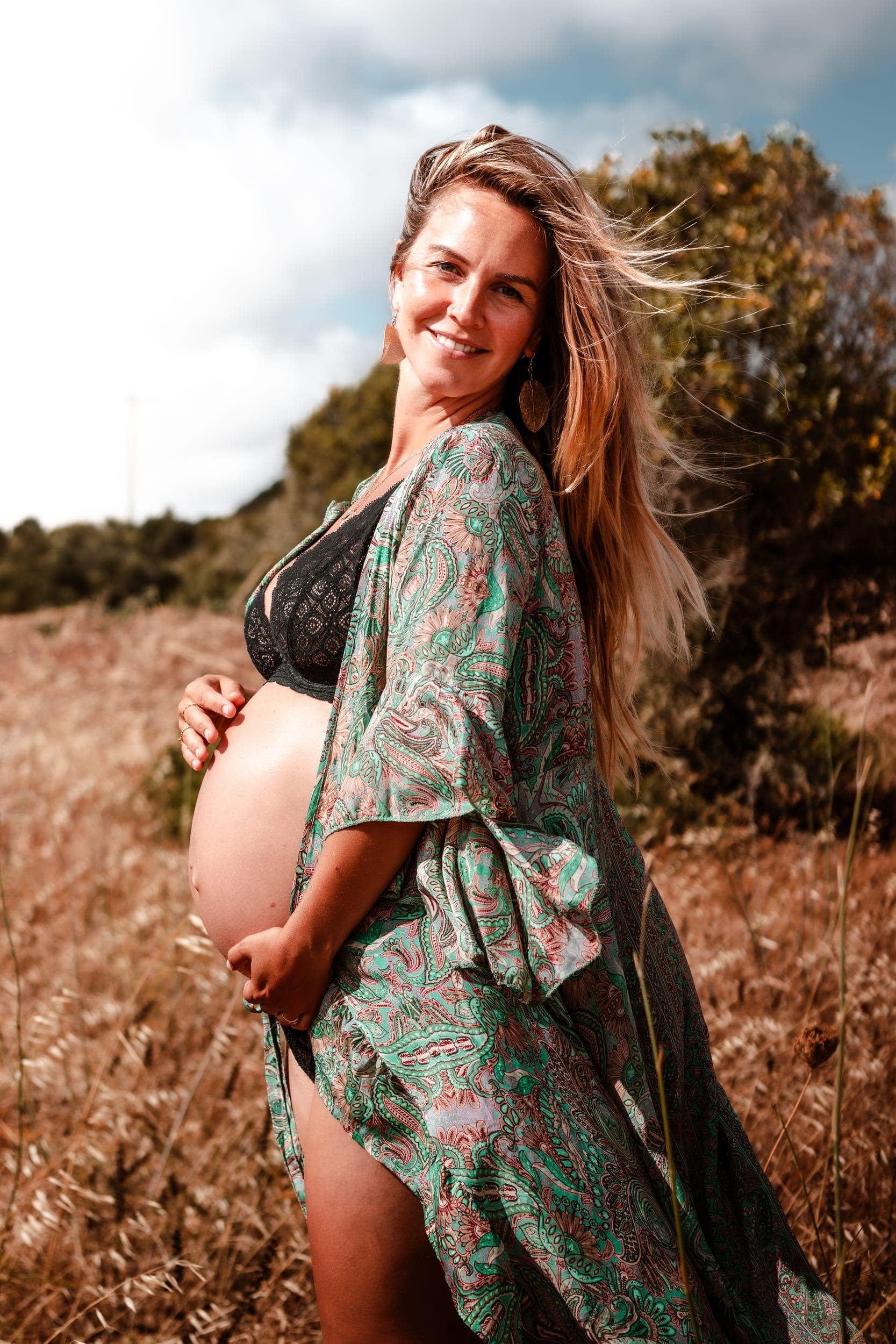 Smiling pregnant woman in paisley robe cradling her bare belly in a golden field
