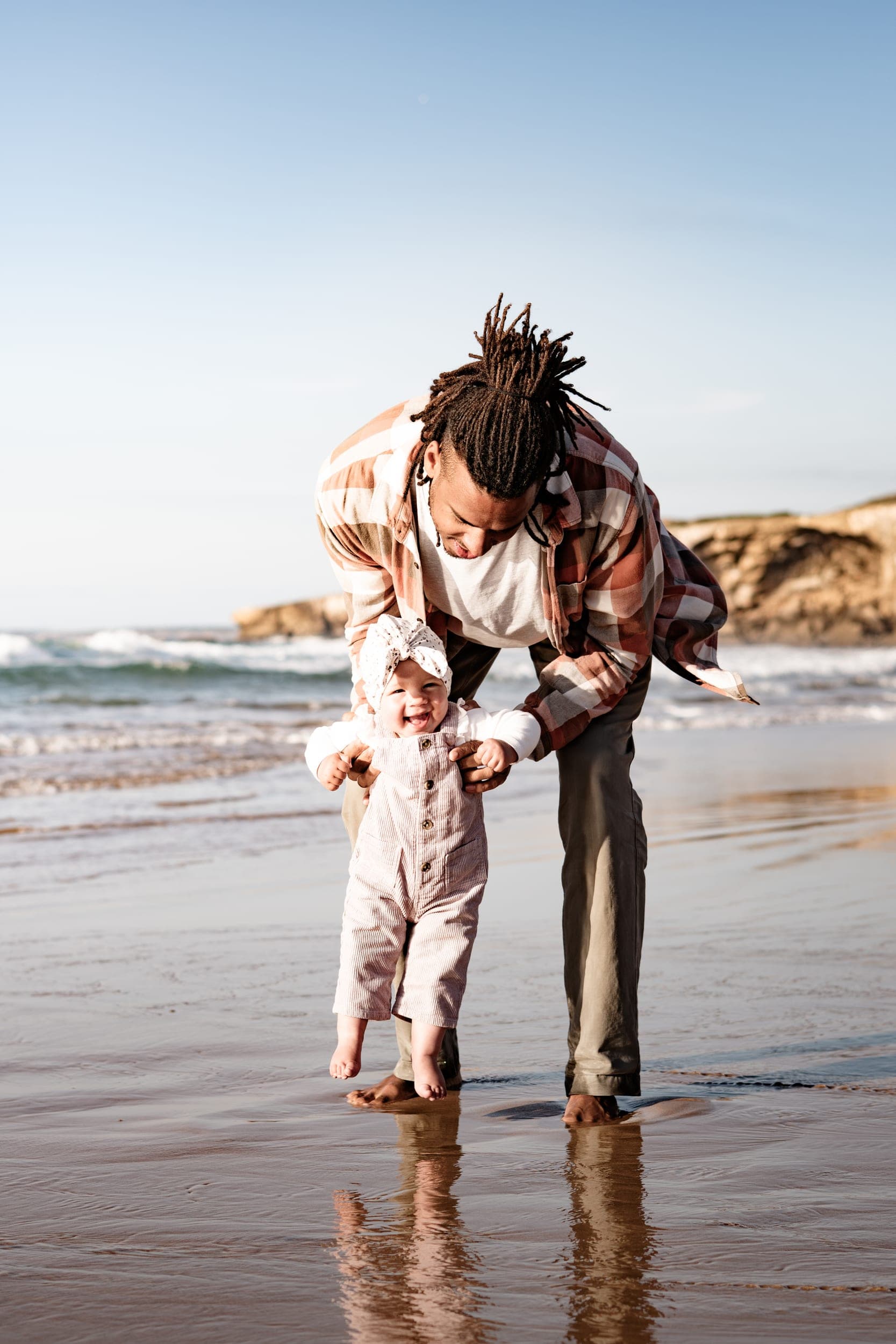 Father helping laughing baby take first steps on a wet Algarve beach at sunset