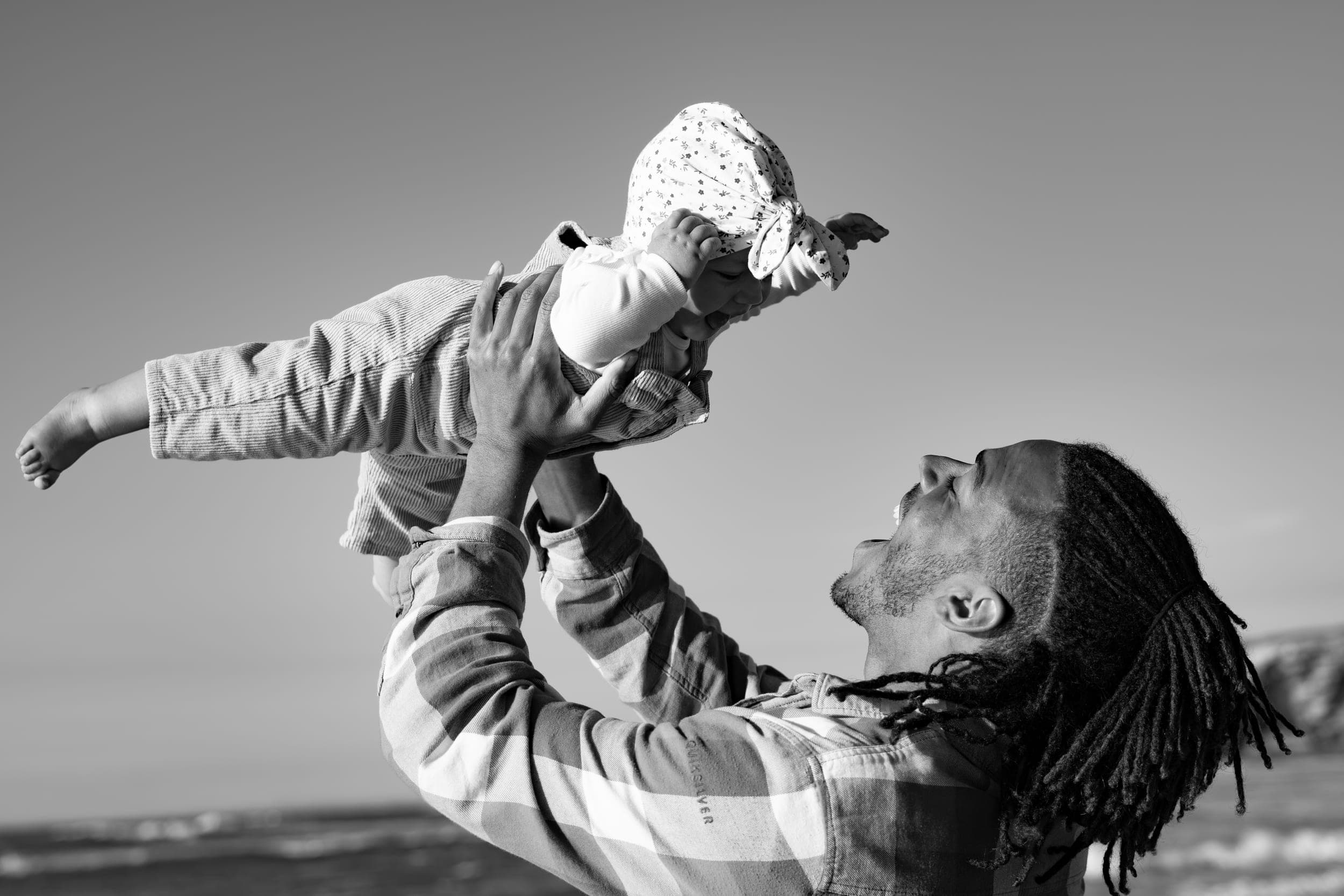 Father lifting laughing baby high into the air on the beach, black and white