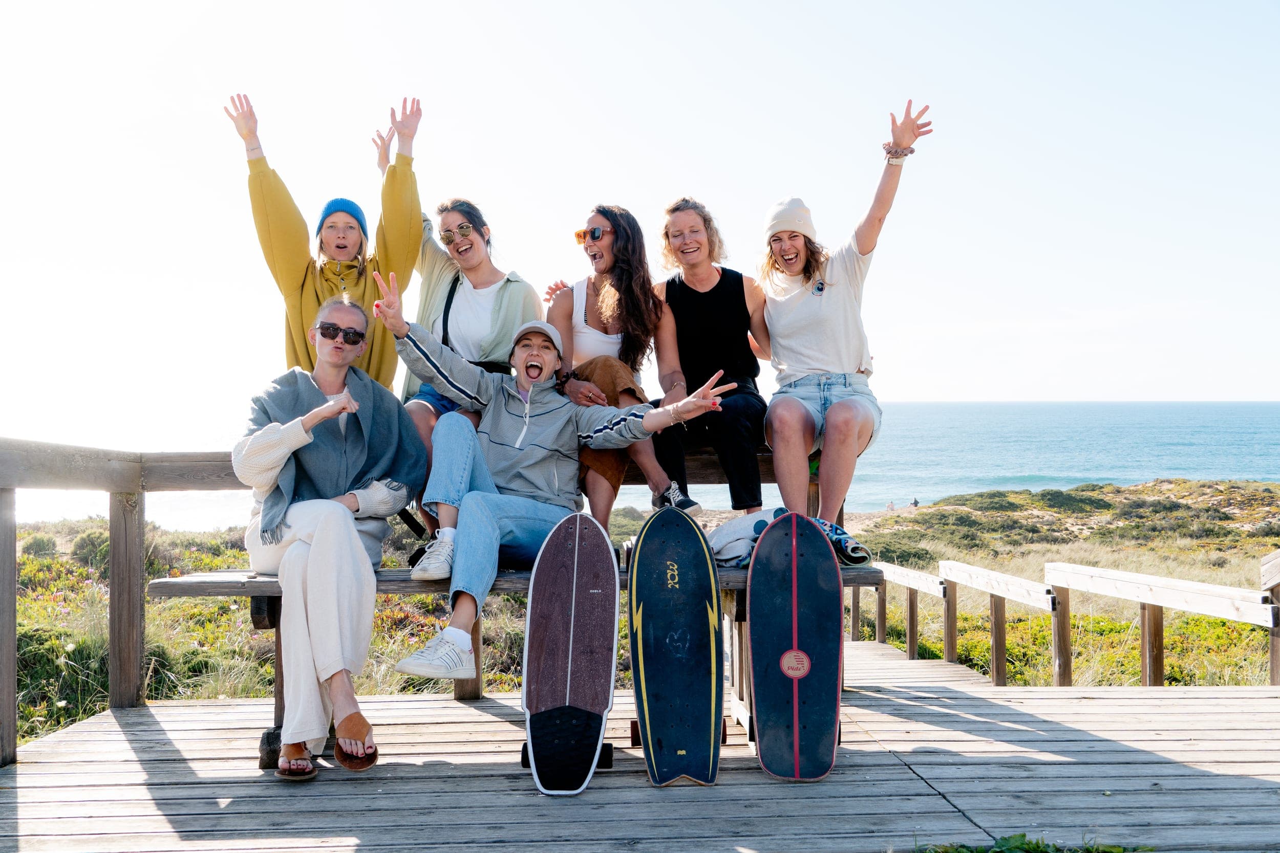 Seven women cheering on a boardwalk bench with surfskate boards, ocean behind them