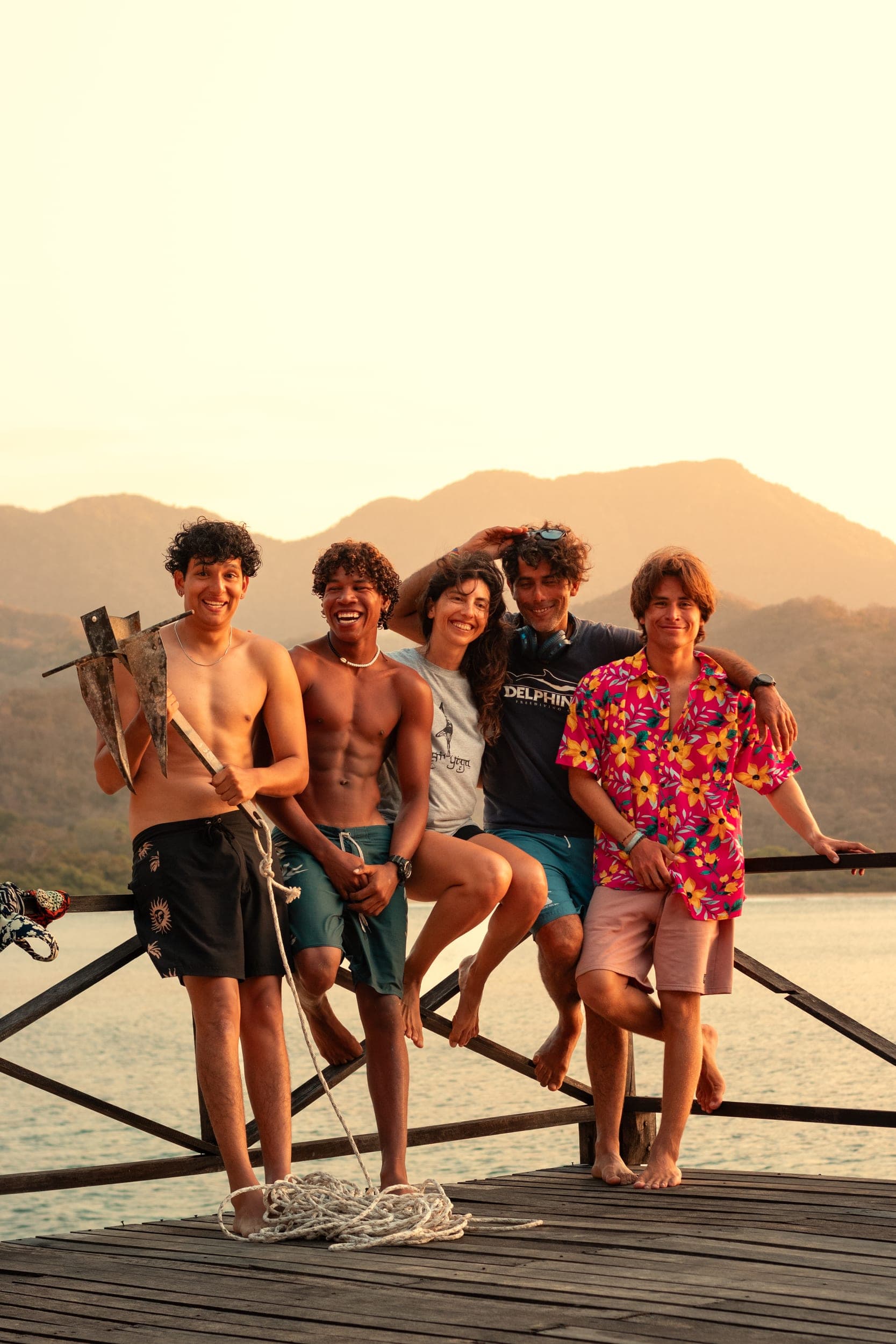 Five friends laughing together on a wooden jetty at golden sunset by calm water and hills