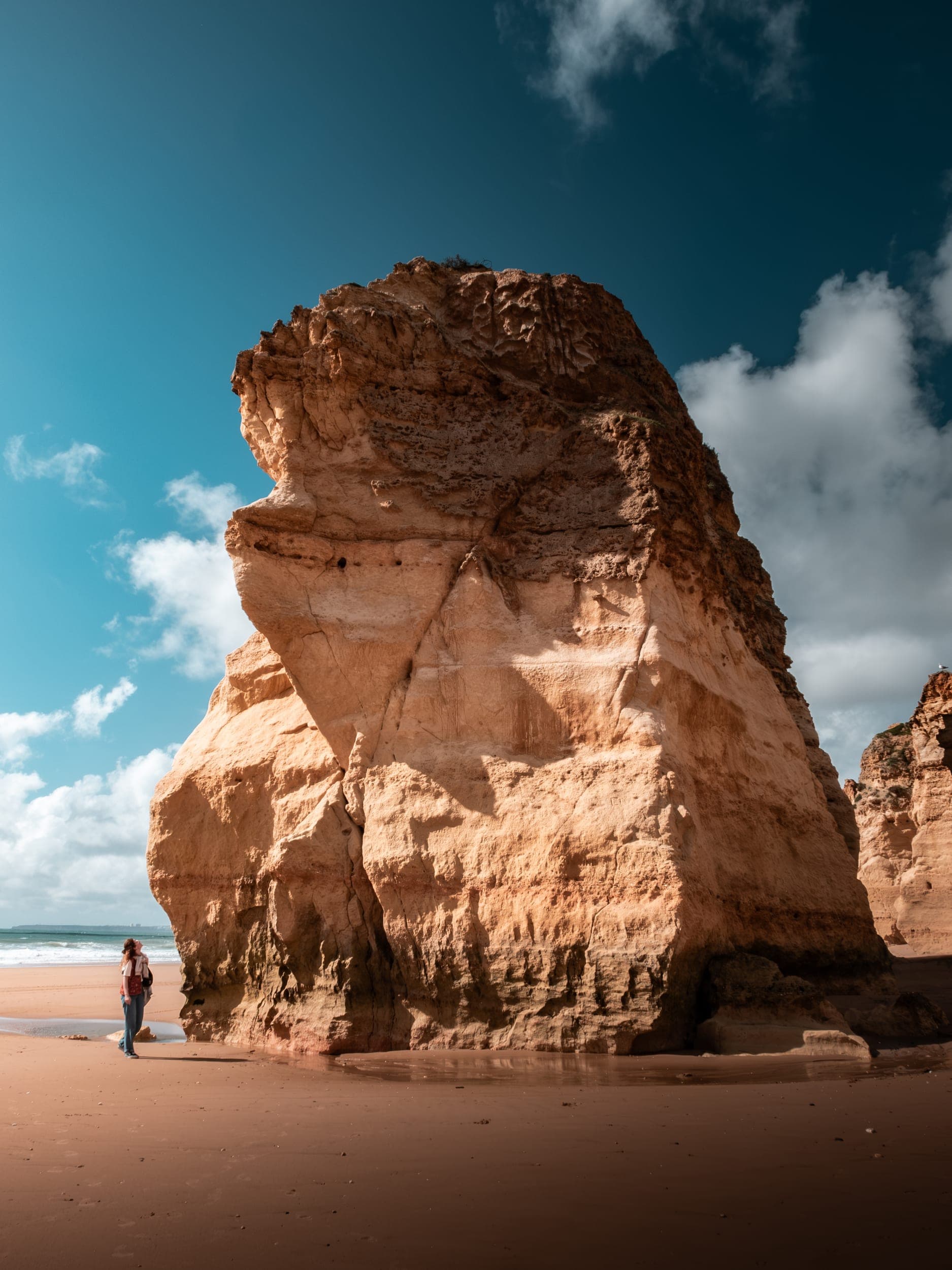 Lone woman standing beside a towering golden cliff on a wide sandy Algarve beach