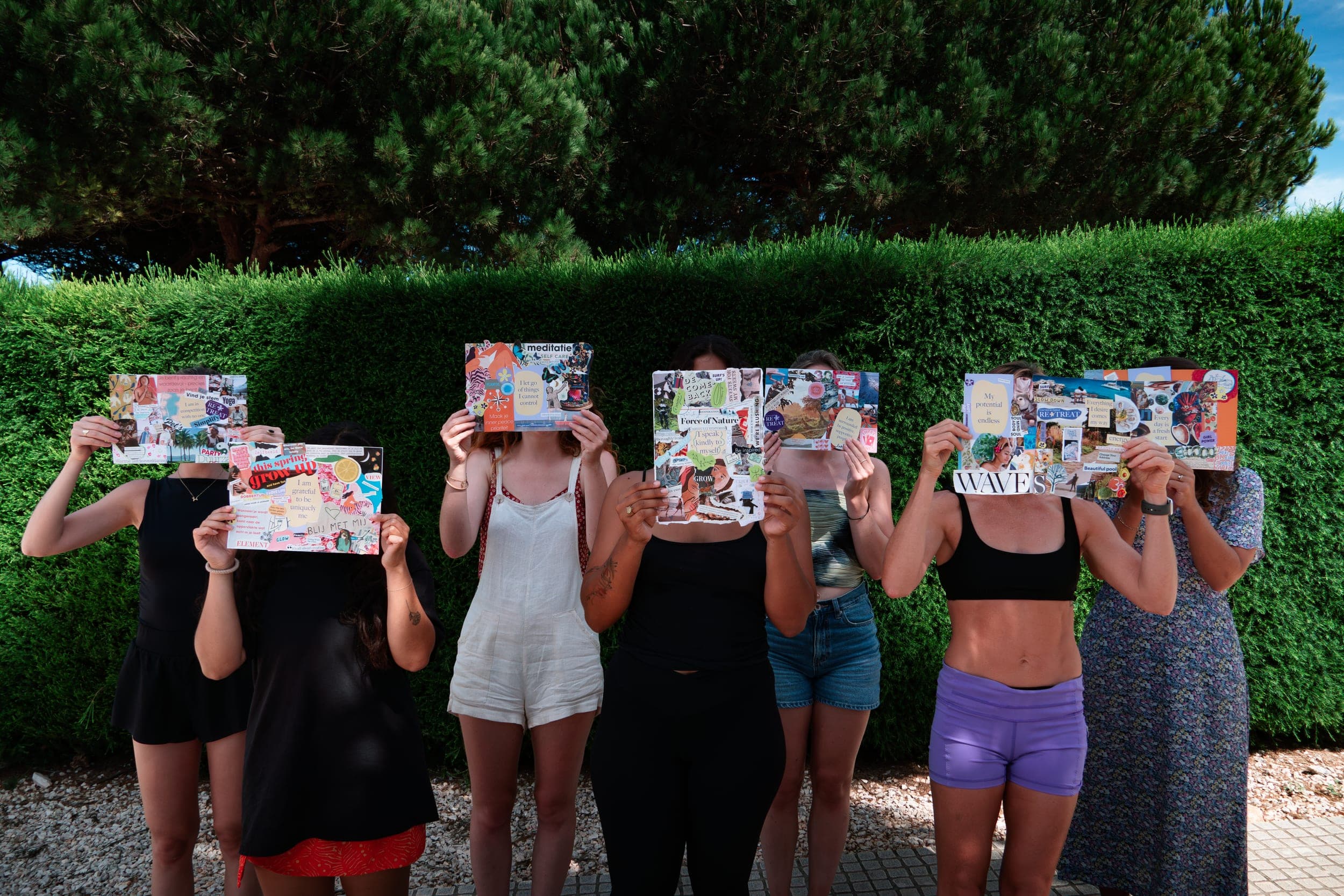 Row of women holding vision-board collages over their faces in front of a green hedge