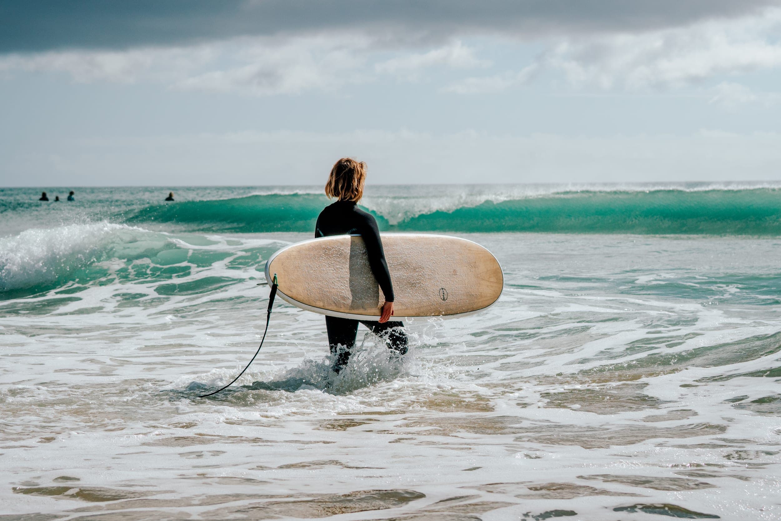 Woman in a wetsuit wading into the surf carrying a wooden longboard toward a breaking wave