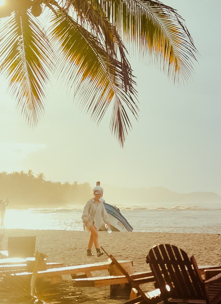 Woman walking on the sand past a beached boat under a palm frond in hazy golden light