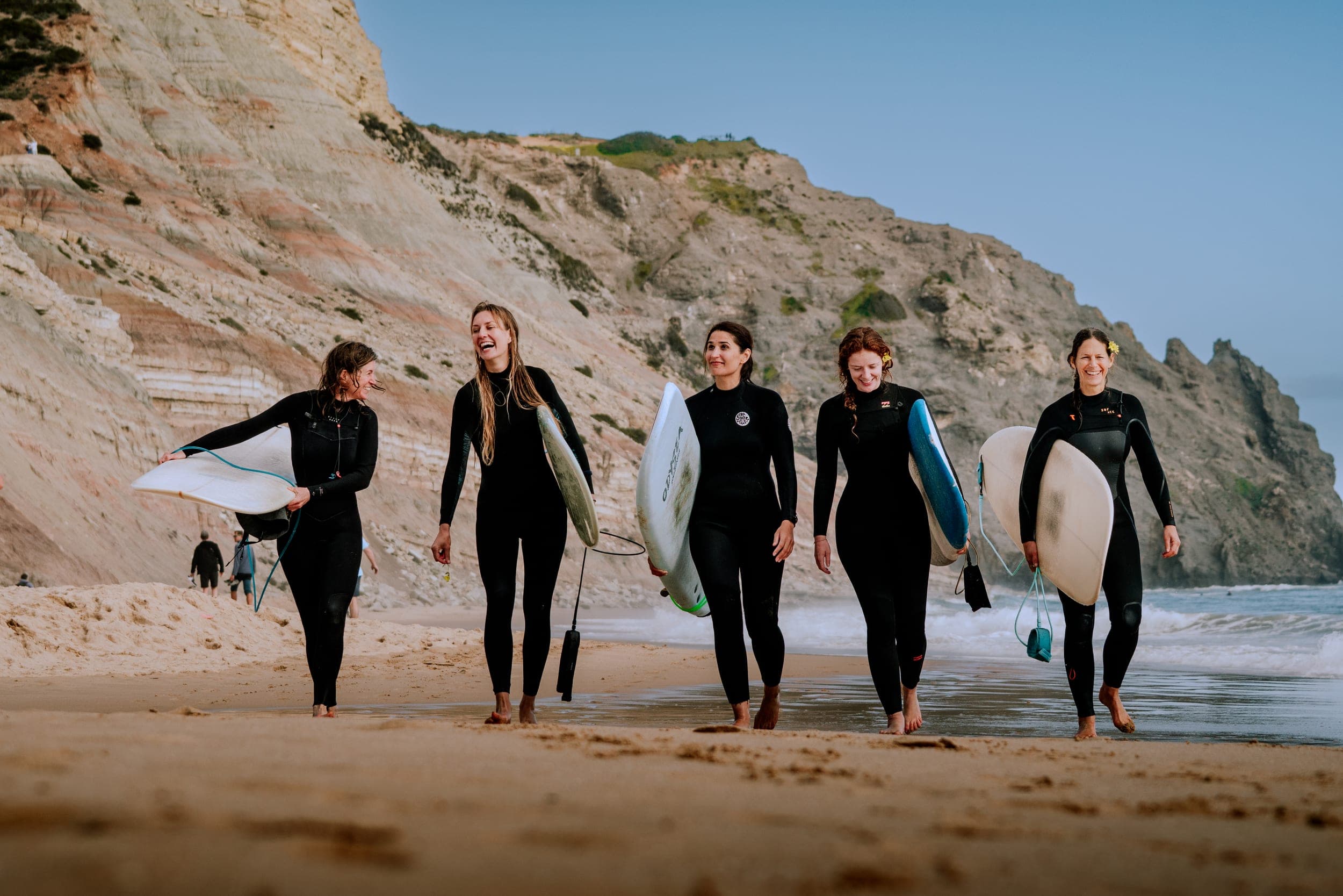 Five smiling women in wetsuits carrying surfboards along the shore beneath tall Algarve cliffs