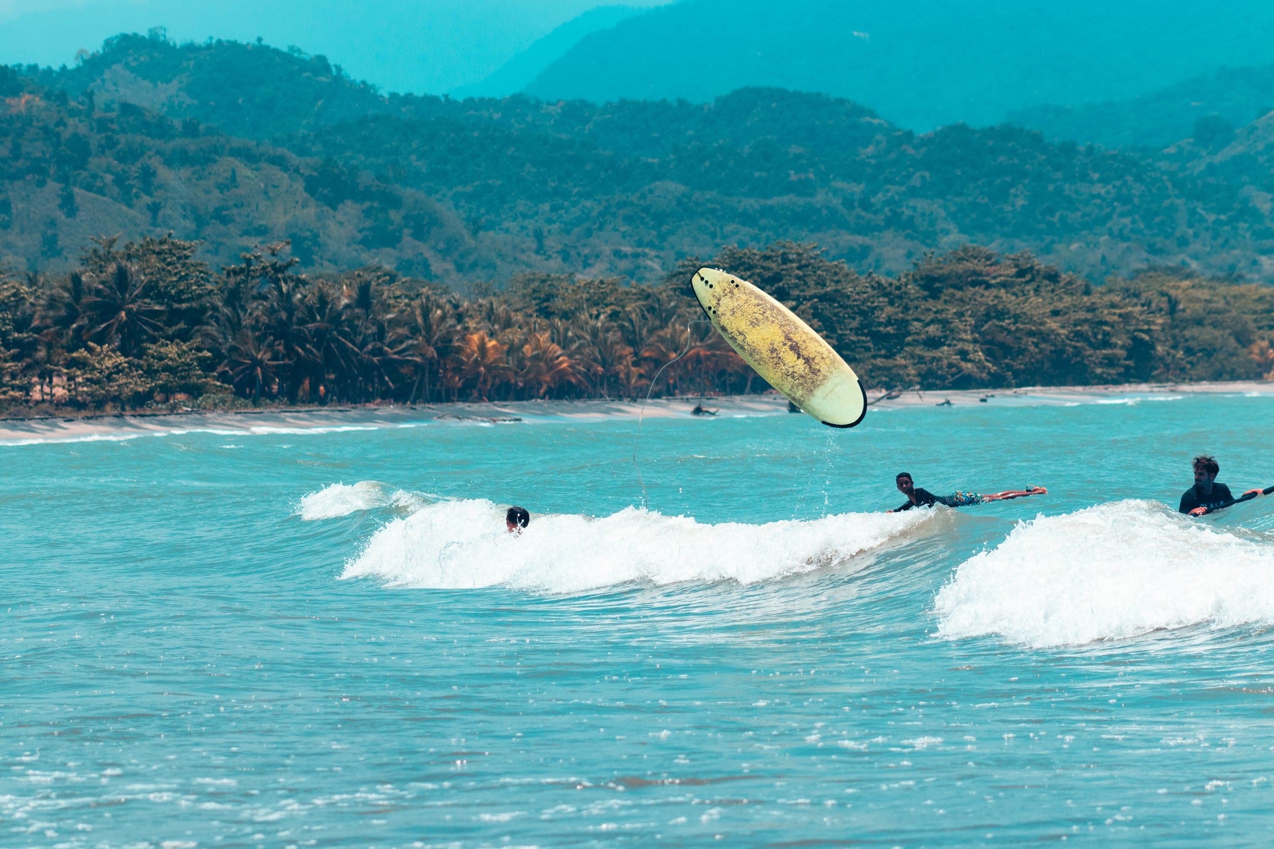 Surfers wiping out as a yellow board flips into the air over turquoise water backed by jungle hills