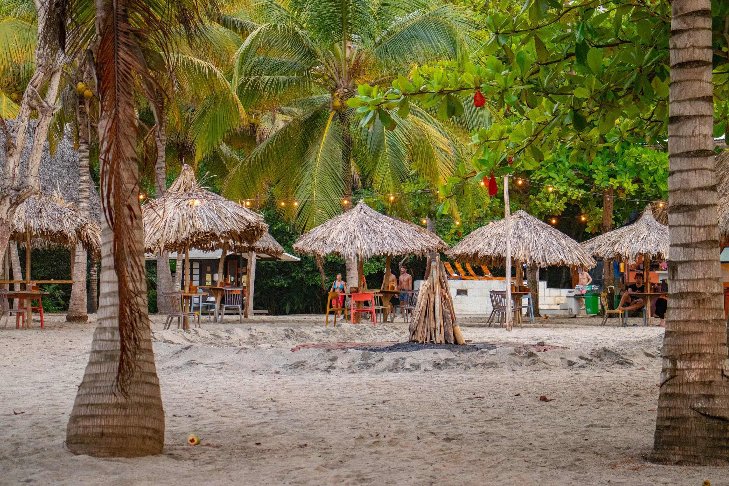 Beach bar with thatched umbrellas and an unlit bonfire among palm trees at dusk