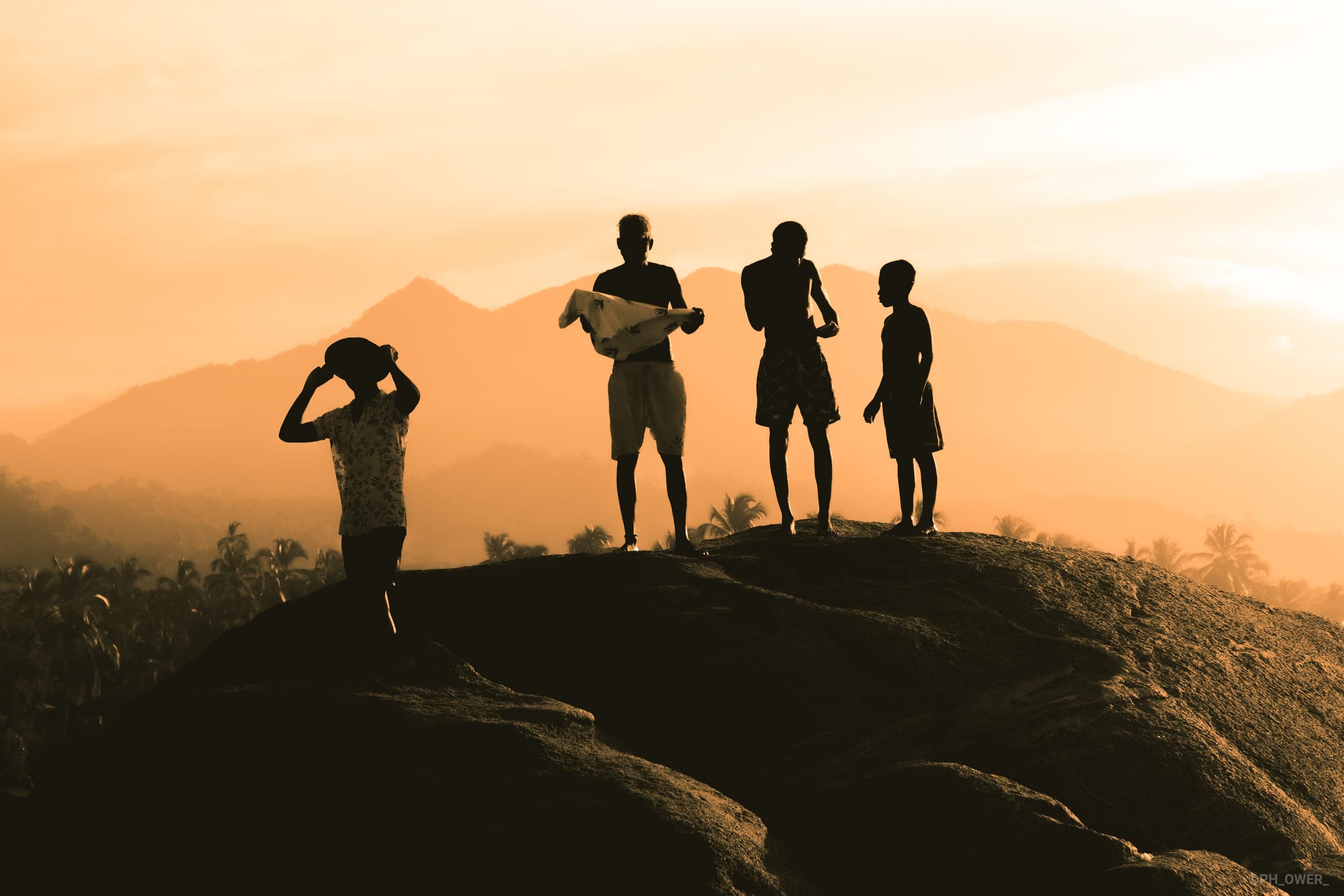 Four people in silhouette standing on a rock at sunset with mountains and palms behind them