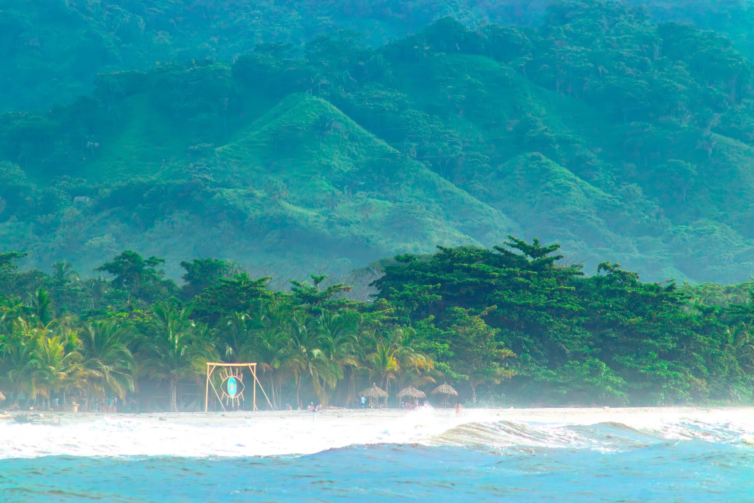 Palm-fringed tropical beach with an eye artwork backed by lush green jungle mountains