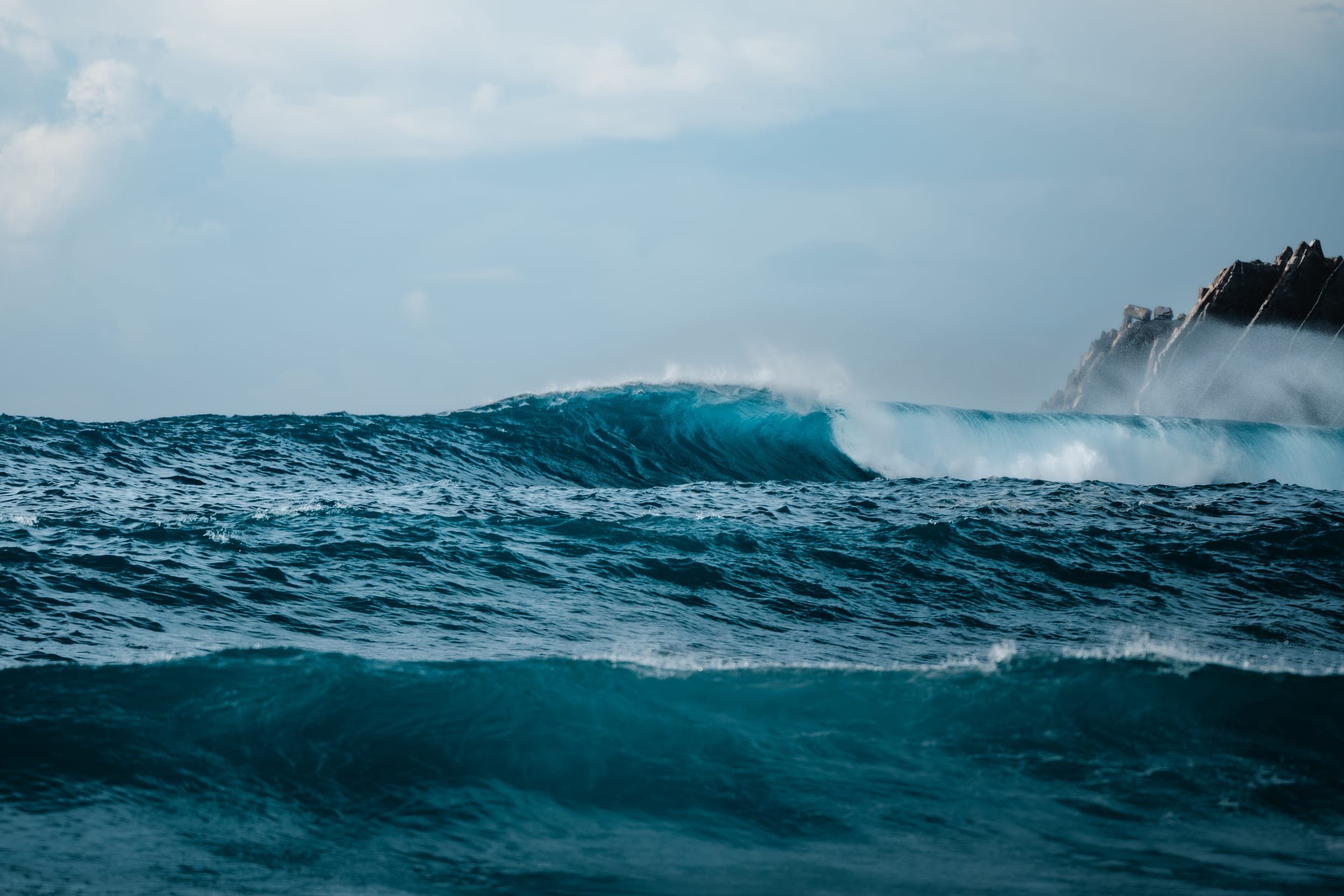 Turquoise wave peeling toward jagged coastal rocks under a soft cloudy sky