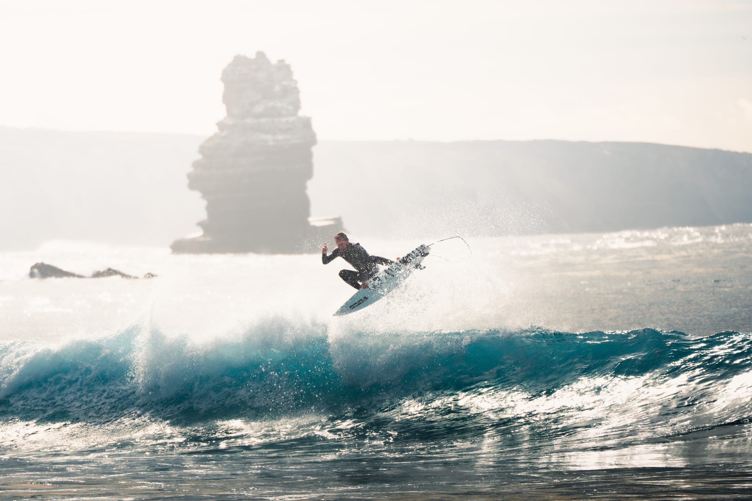 Surfer launching an aerial off a wave with a rugged rock stack behind in backlit spray