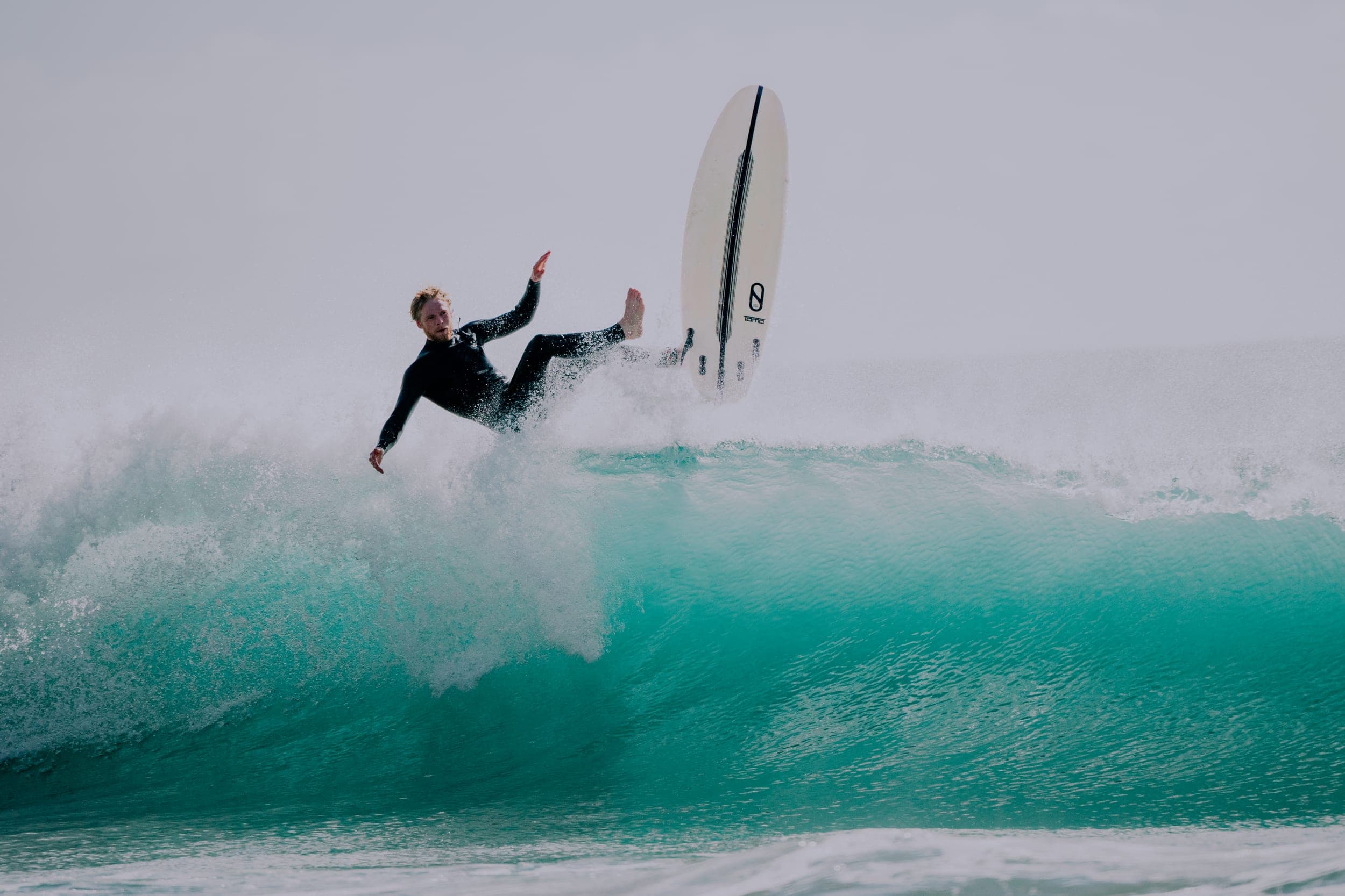 Surfer in a black wetsuit falling off his board on a breaking turquoise wave
