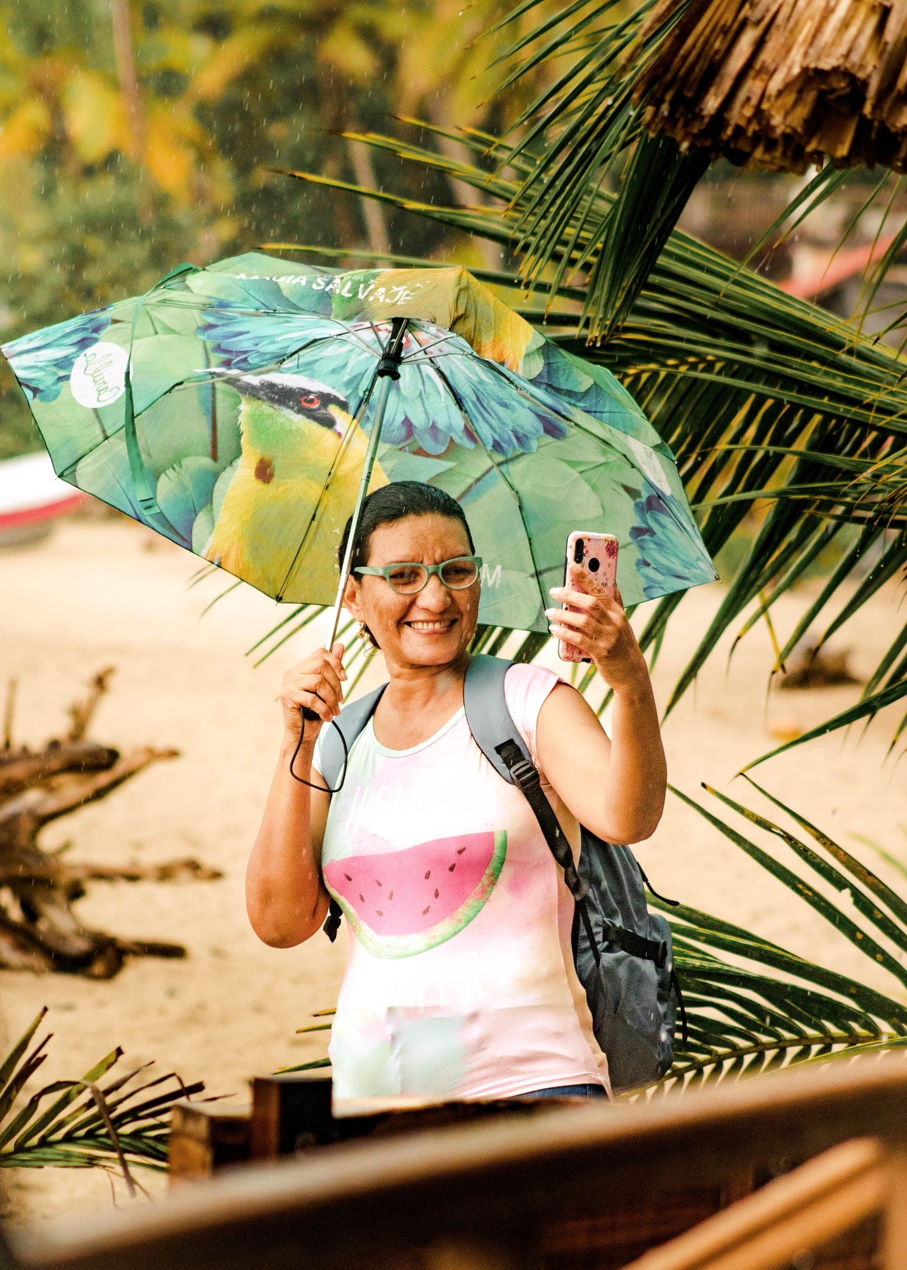 Smiling woman with a parrot-print umbrella taking a selfie in the rain on a tropical beach