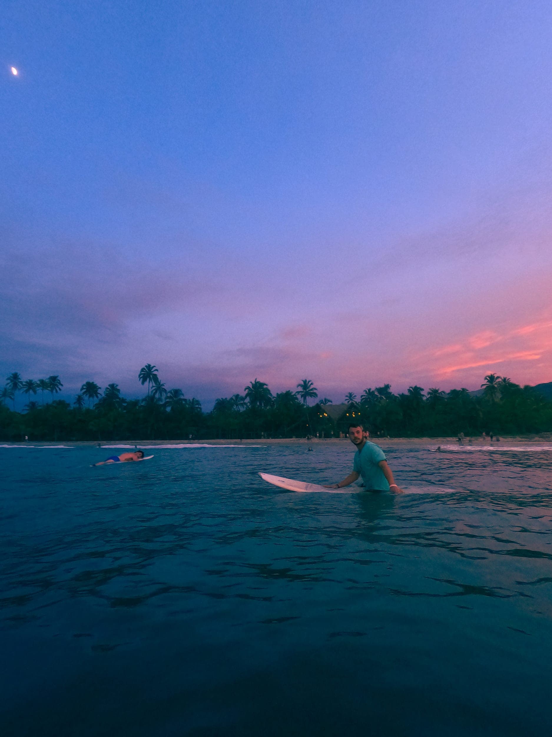 Two surfers in the sea at dusk, palm-lined shore glowing pink under a crescent moon