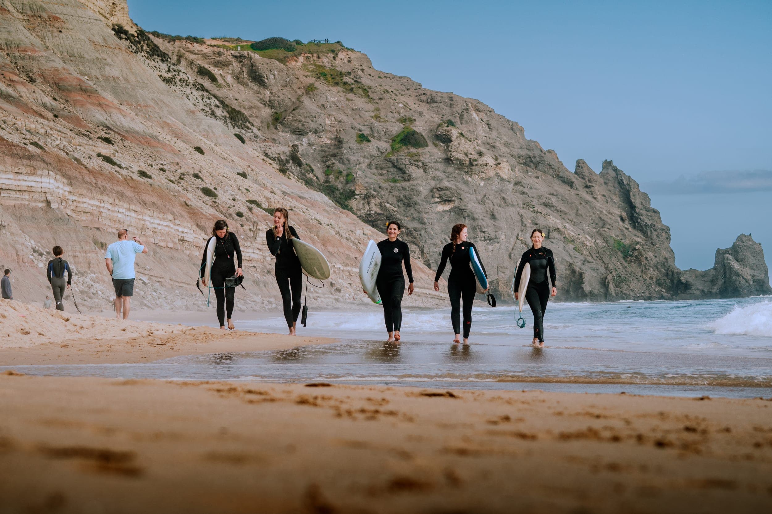 Five women in wetsuits carrying surfboards along the shoreline beneath towering Algarve cliffs