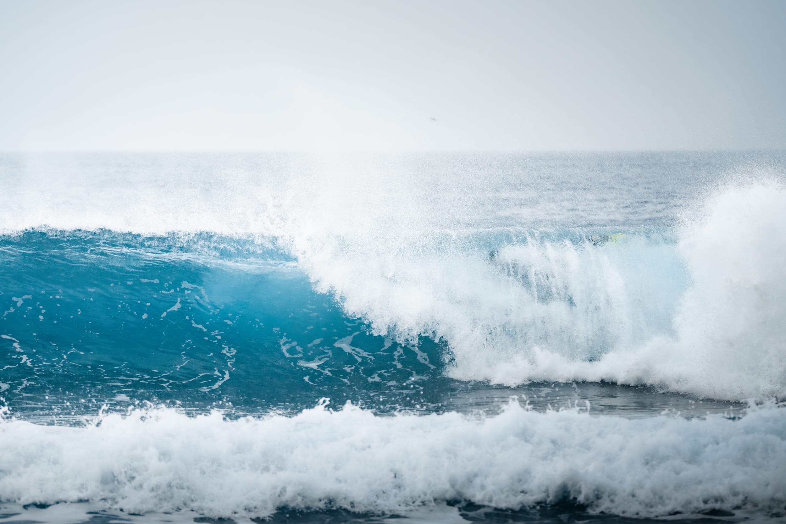Powerful blue wave breaking into white spray under a pale hazy sky