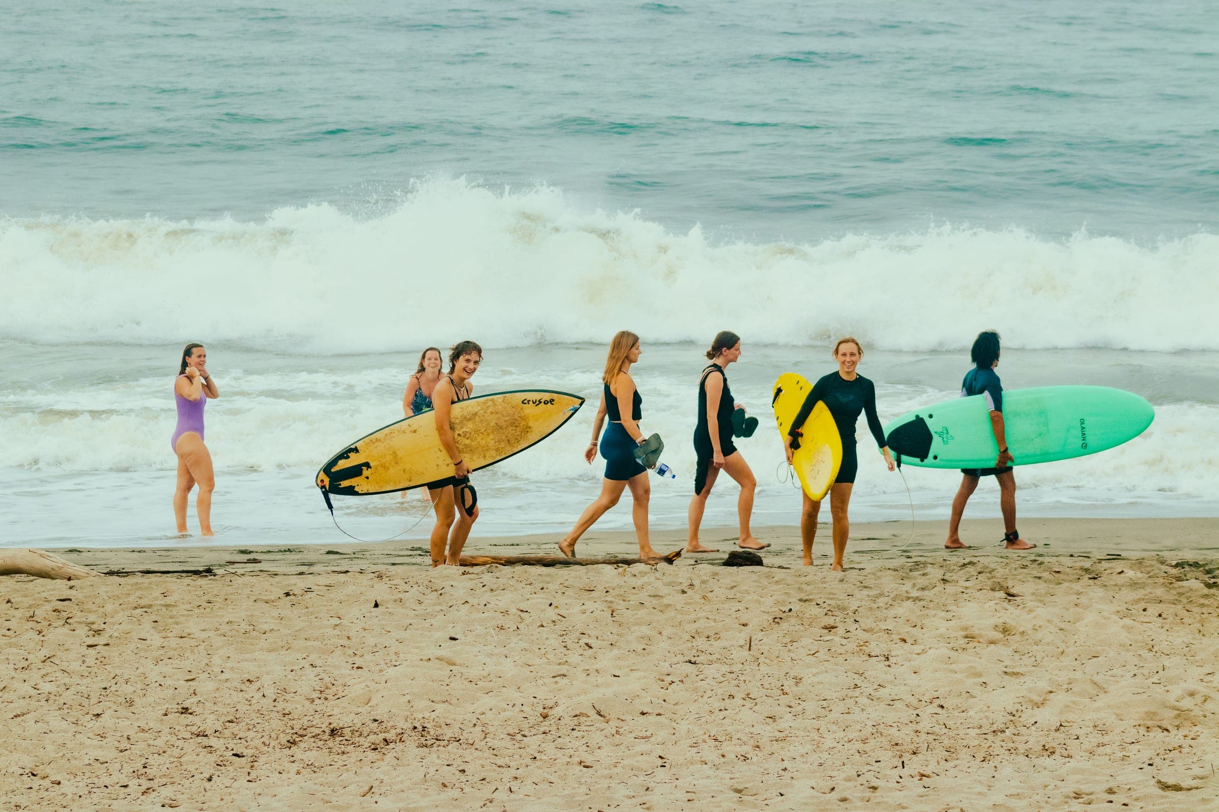Group of women in swimwear walking along the sand carrying colourful surfboards past breaking waves