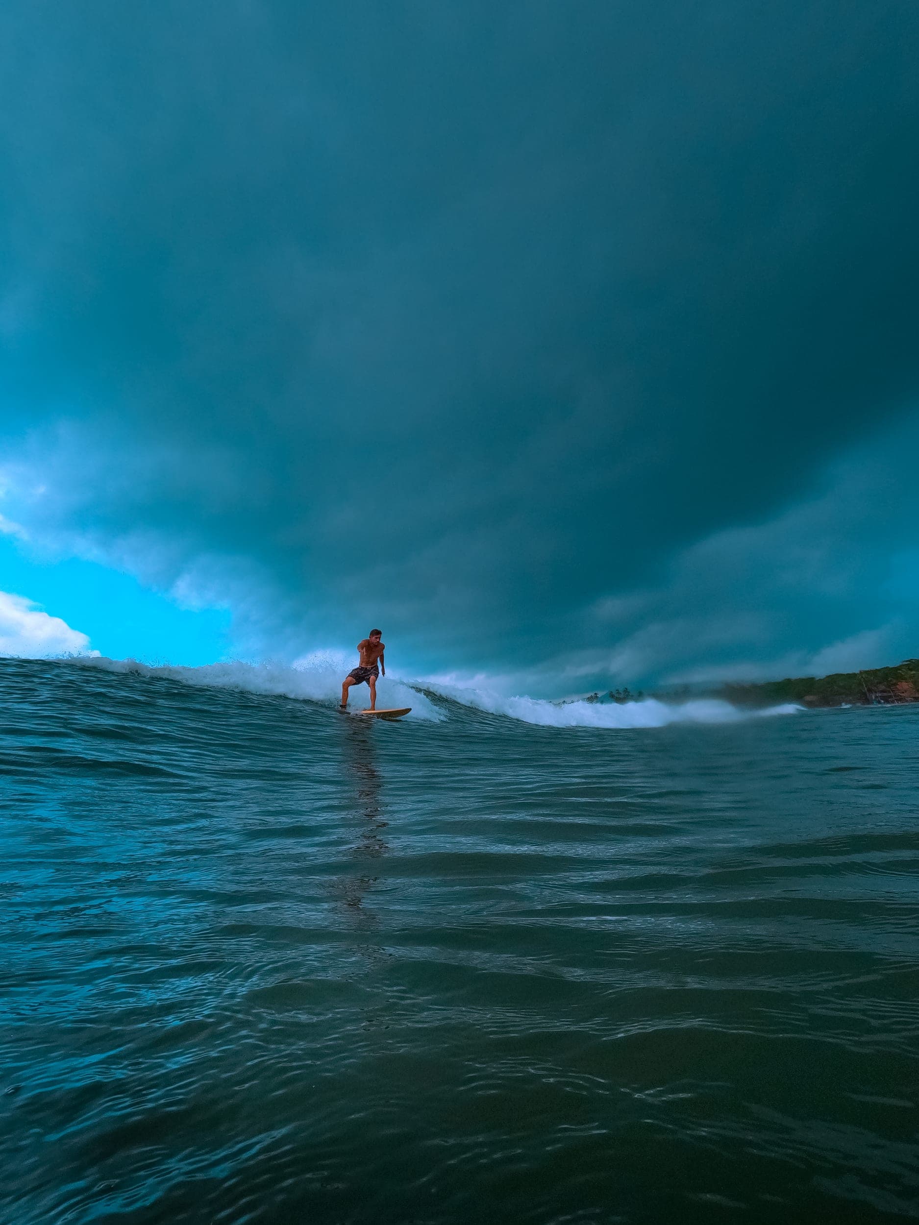 Shirtless surfer riding a small wave on a longboard under a moody teal stormy sky