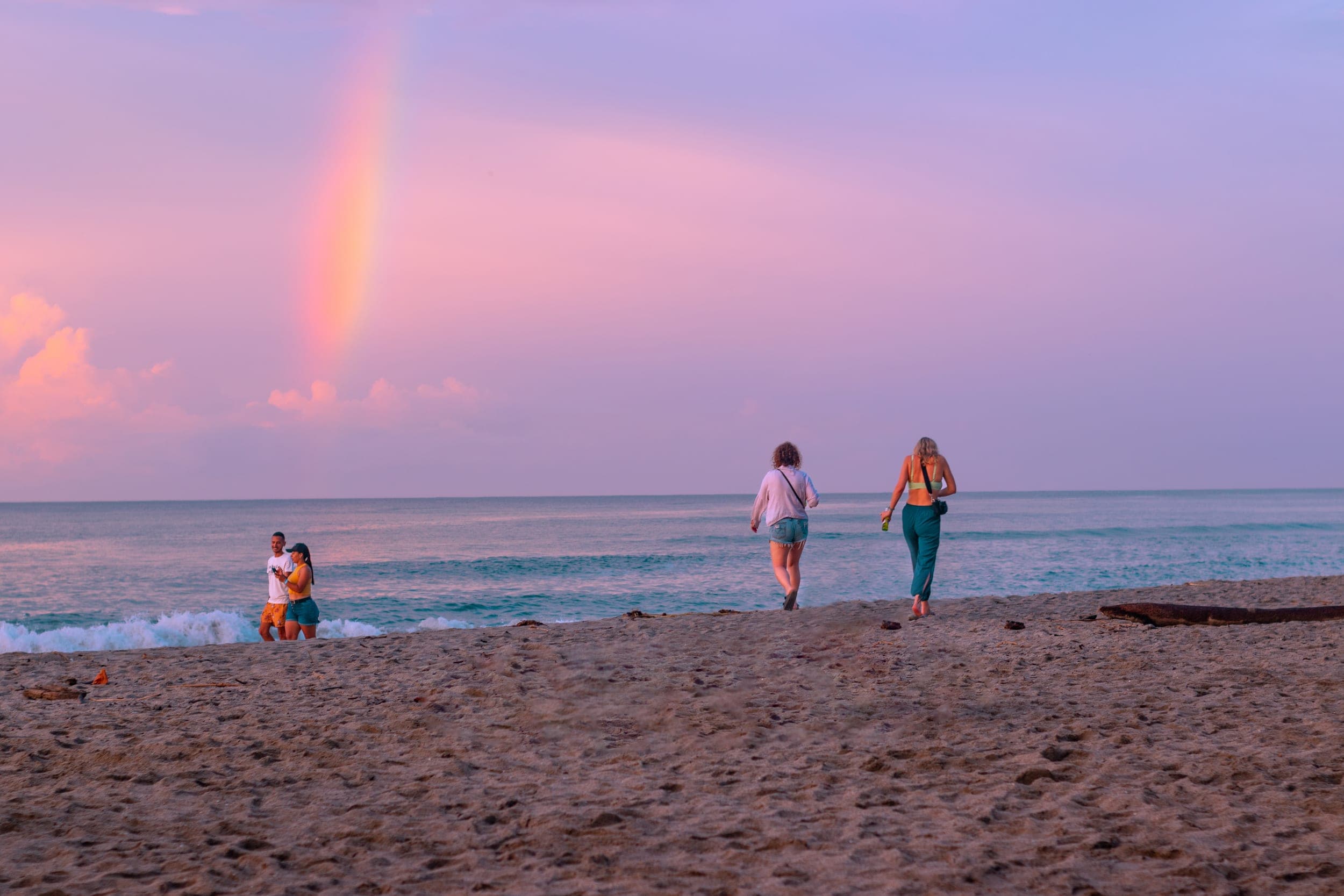 Two women walking a sandy beach toward a couple by the sea under a rainbow at dusk
