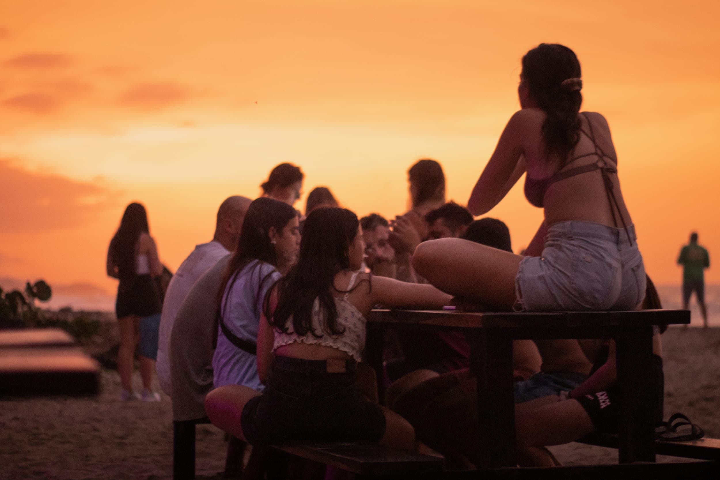 Young people gathered around a wooden table on the beach at a glowing orange sunset