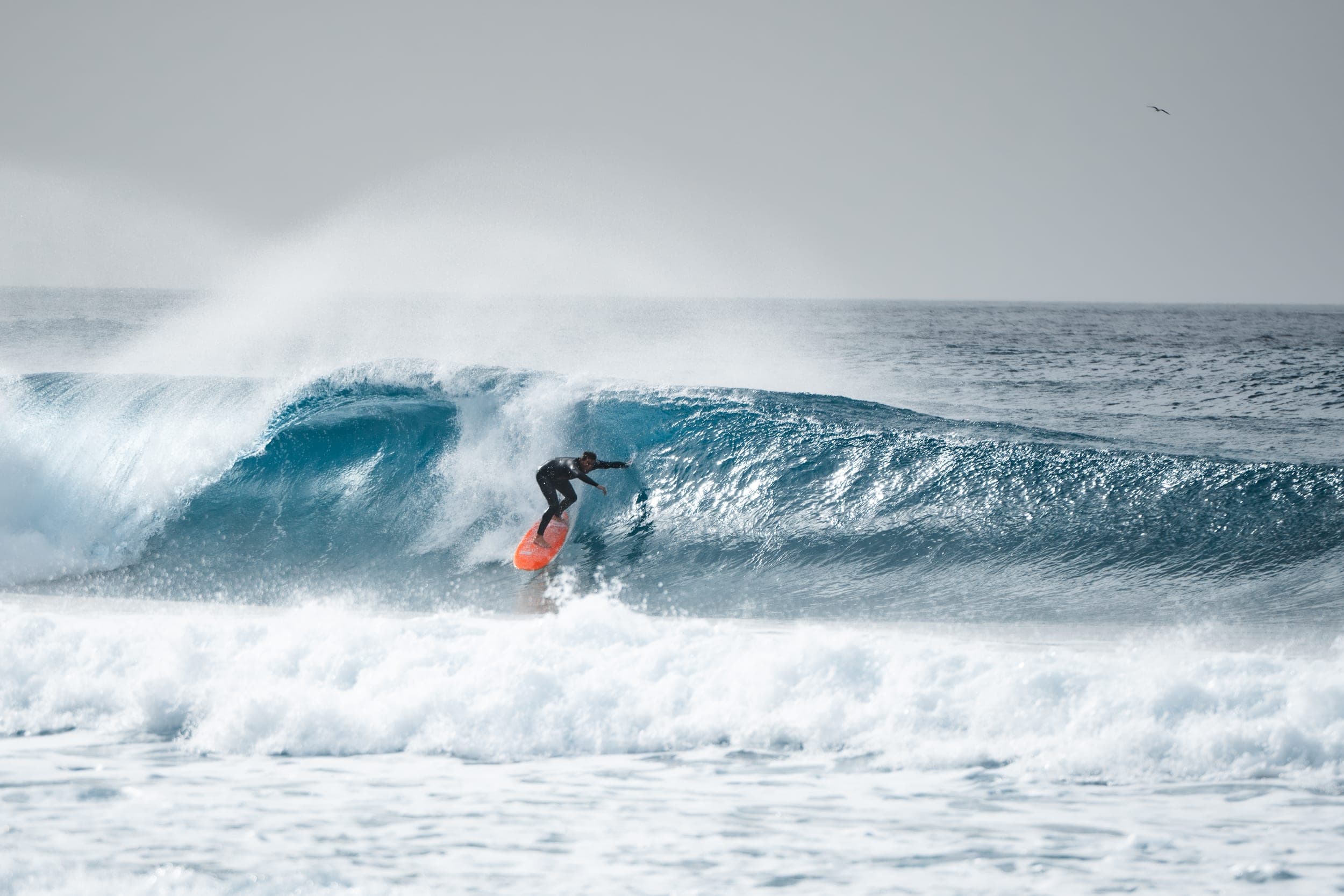 Surfer in a wetsuit riding an orange board across a large peeling blue wave