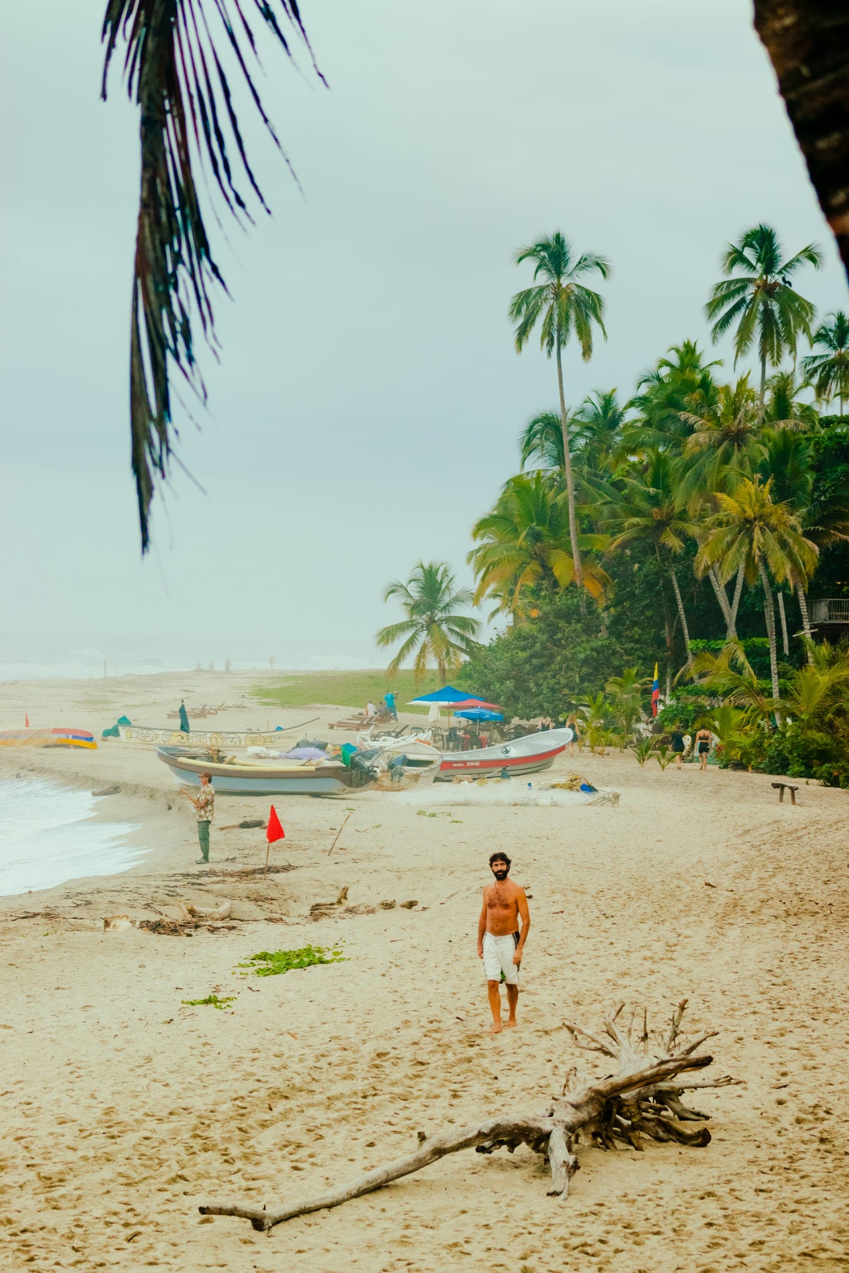 Shirtless man standing on a palm-lined tropical beach with fishing boats and grey sky