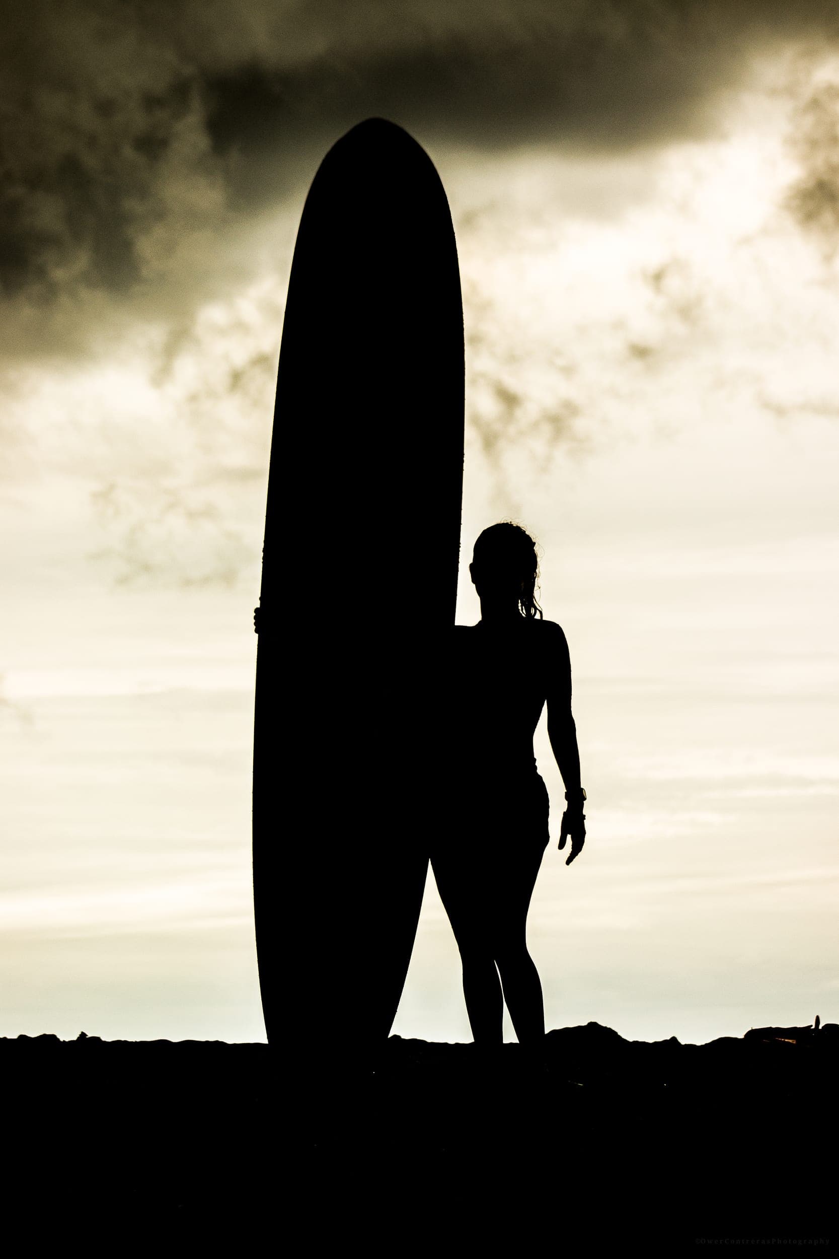 Silhouette of a woman standing beside her longboard against a dramatic cloudy sky