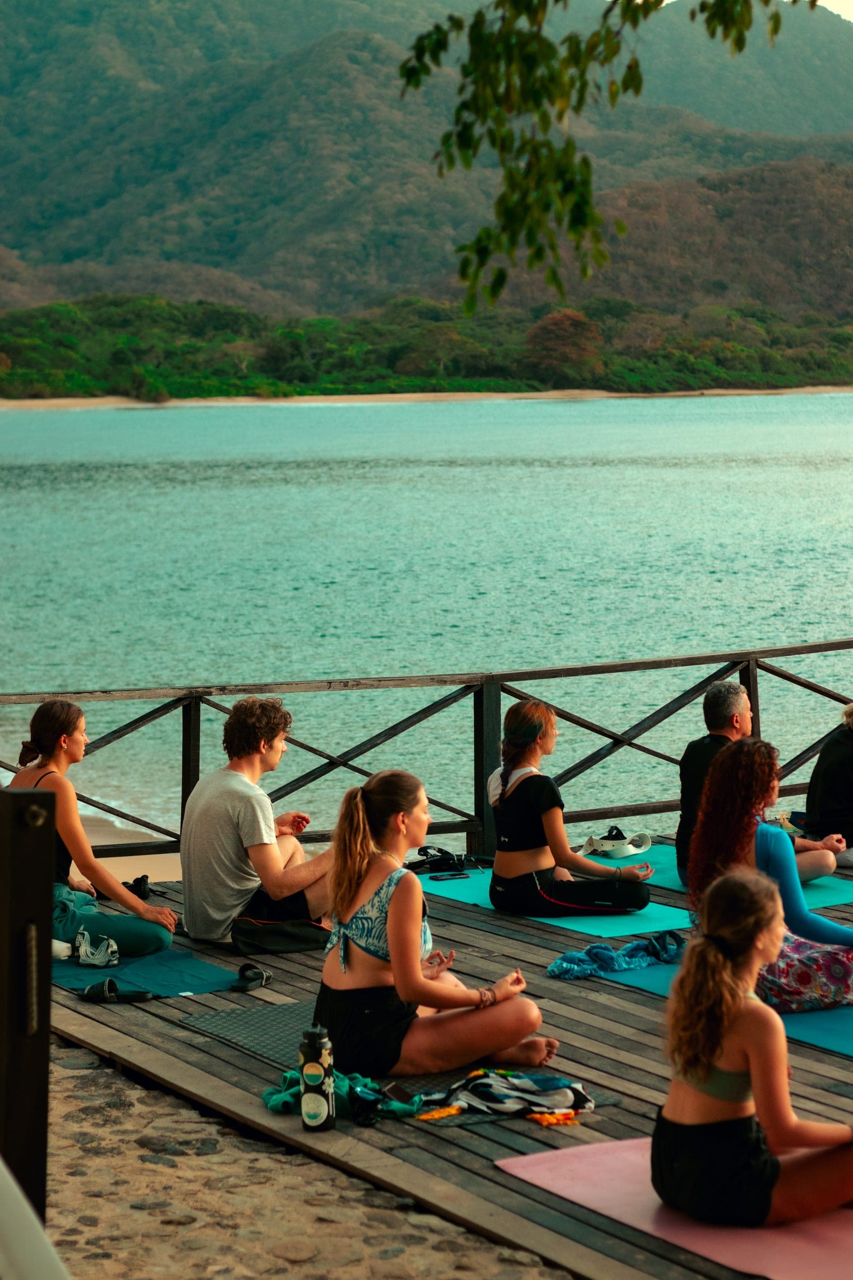 Group meditating on yoga mats on a wooden deck beside a calm bay at dusk