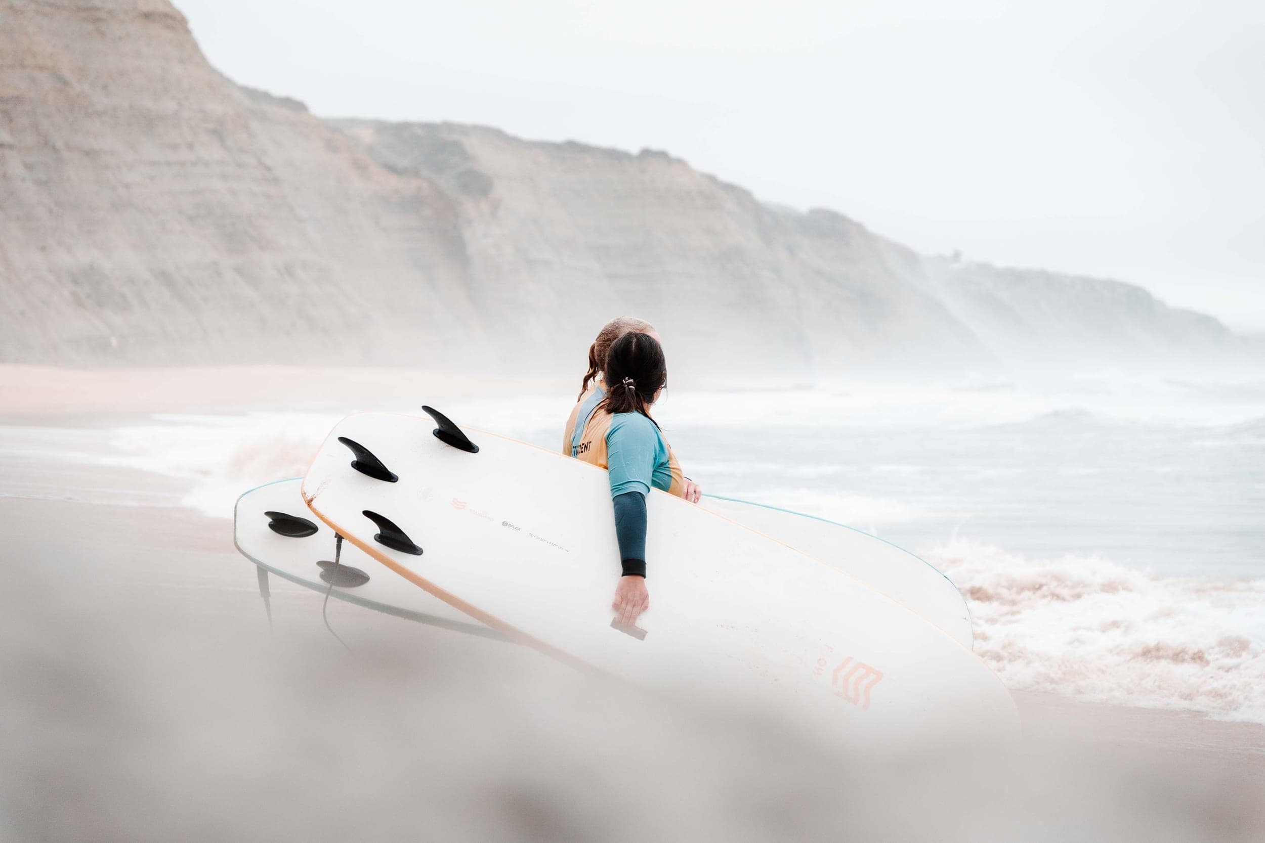 Woman in wetsuit carrying a surfboard toward the misty sea below tall cliffs