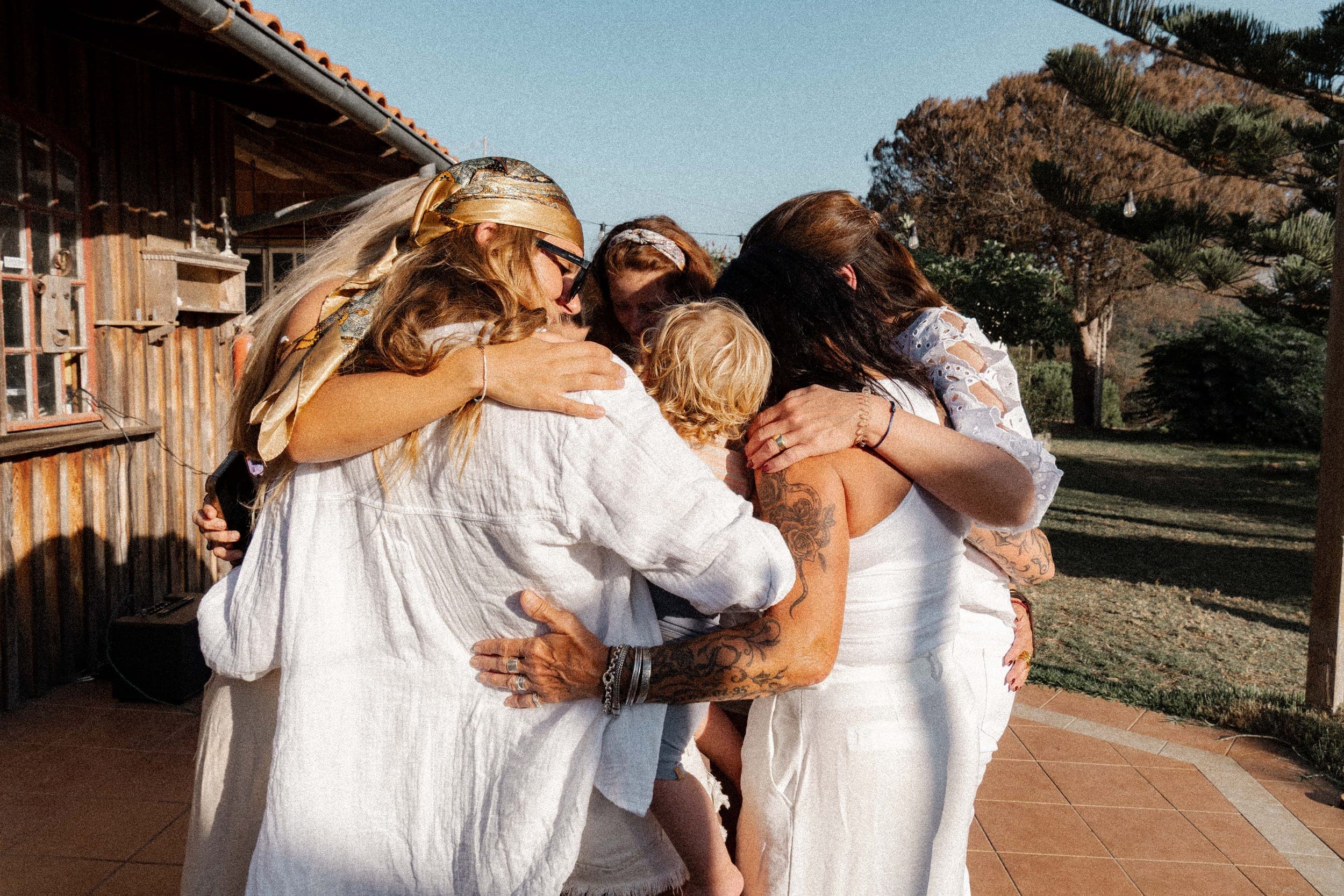 Women and a child sharing a group hug outside a rustic wooden house at golden hour