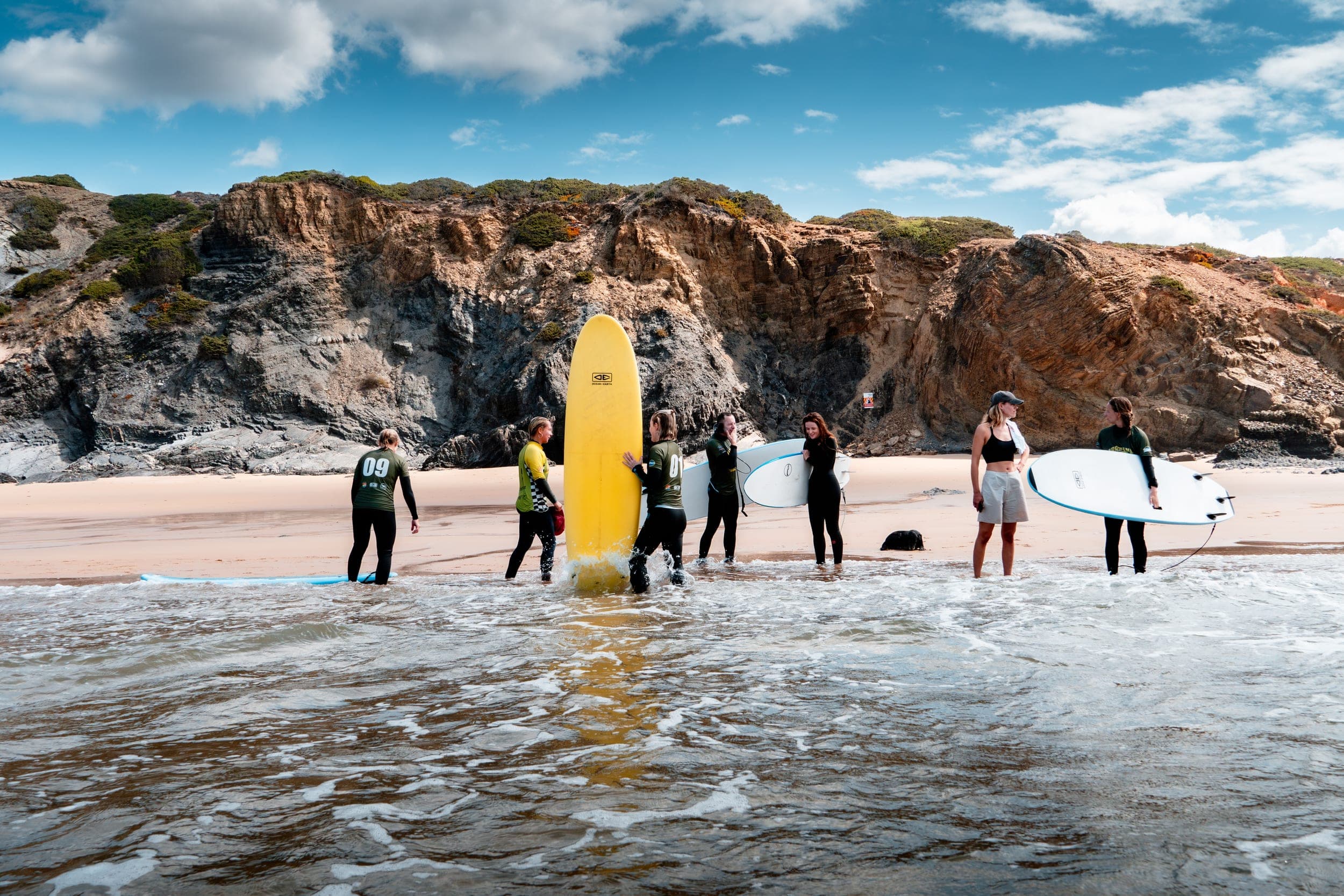 Surf group in wetsuits with boards wading into the shore below rugged cliffs