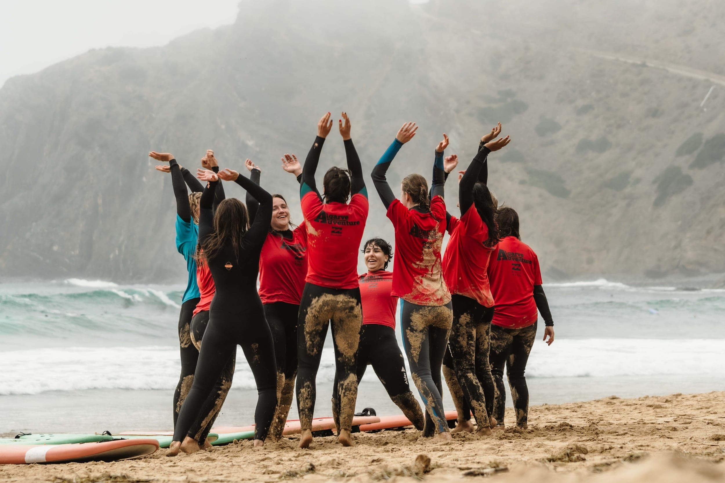 Surf group in wetsuits cheering with raised arms on a misty cliff-backed beach