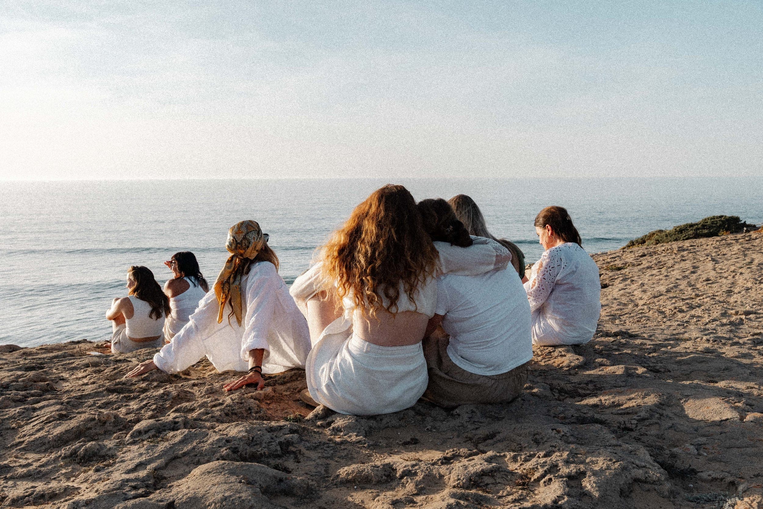 Group of women in white sitting close together watching the sea at sunset