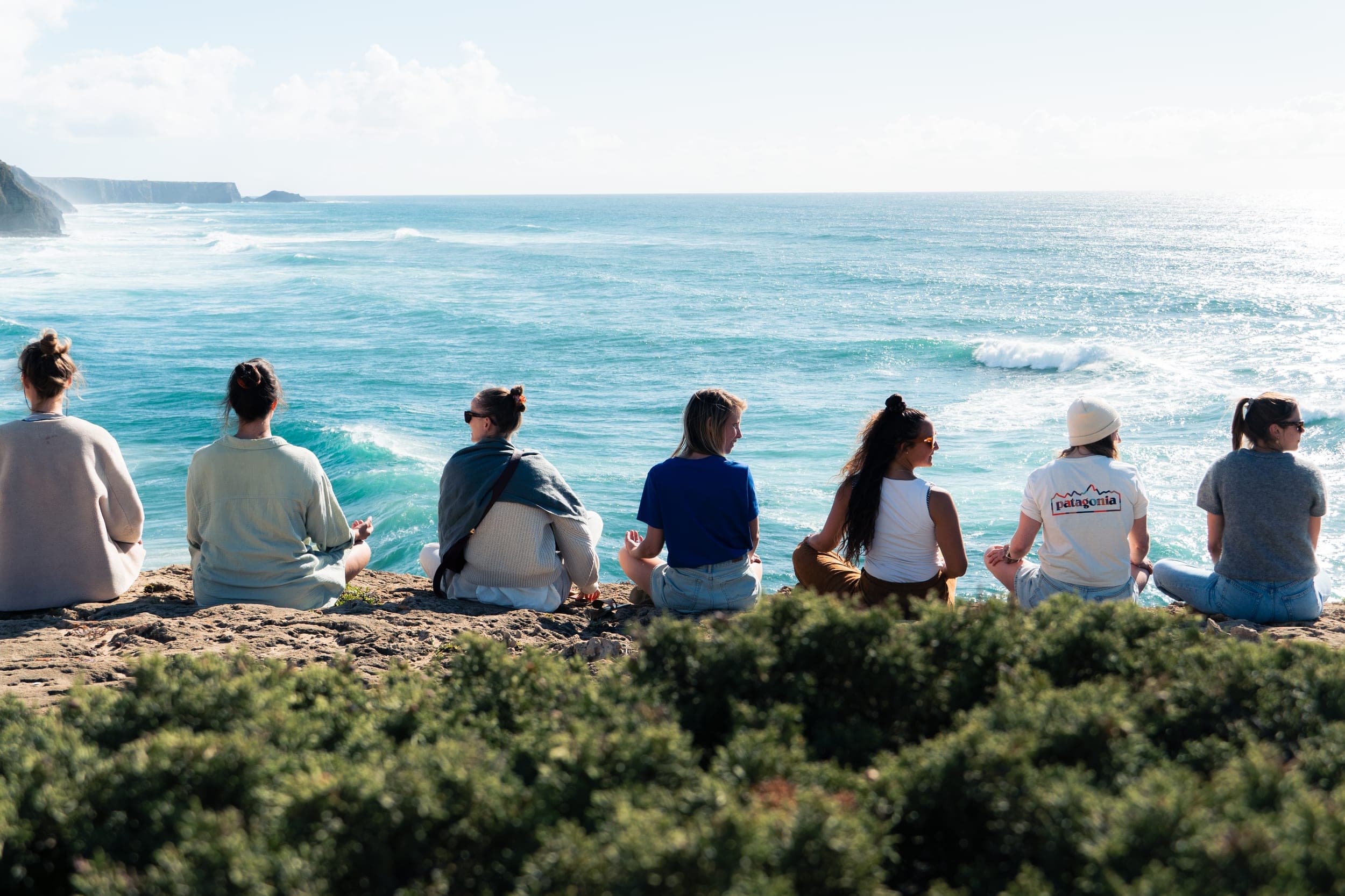 Group of women meditating in a row on a clifftop overlooking the turquoise ocean