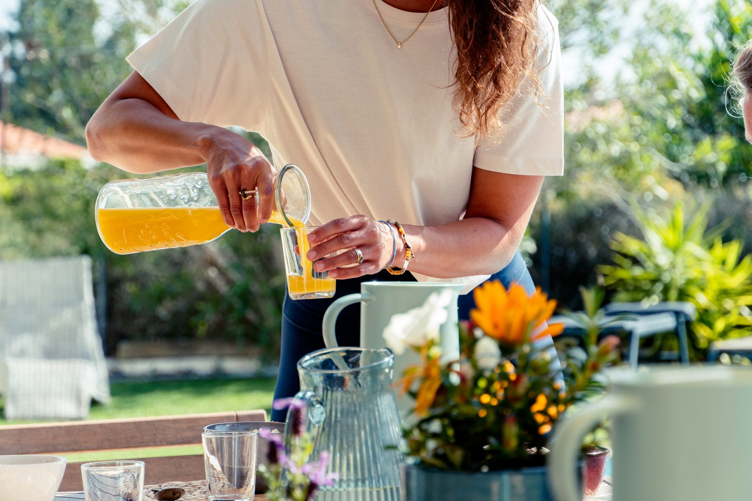 Woman pouring fresh orange juice into a glass at a sunny outdoor breakfast table