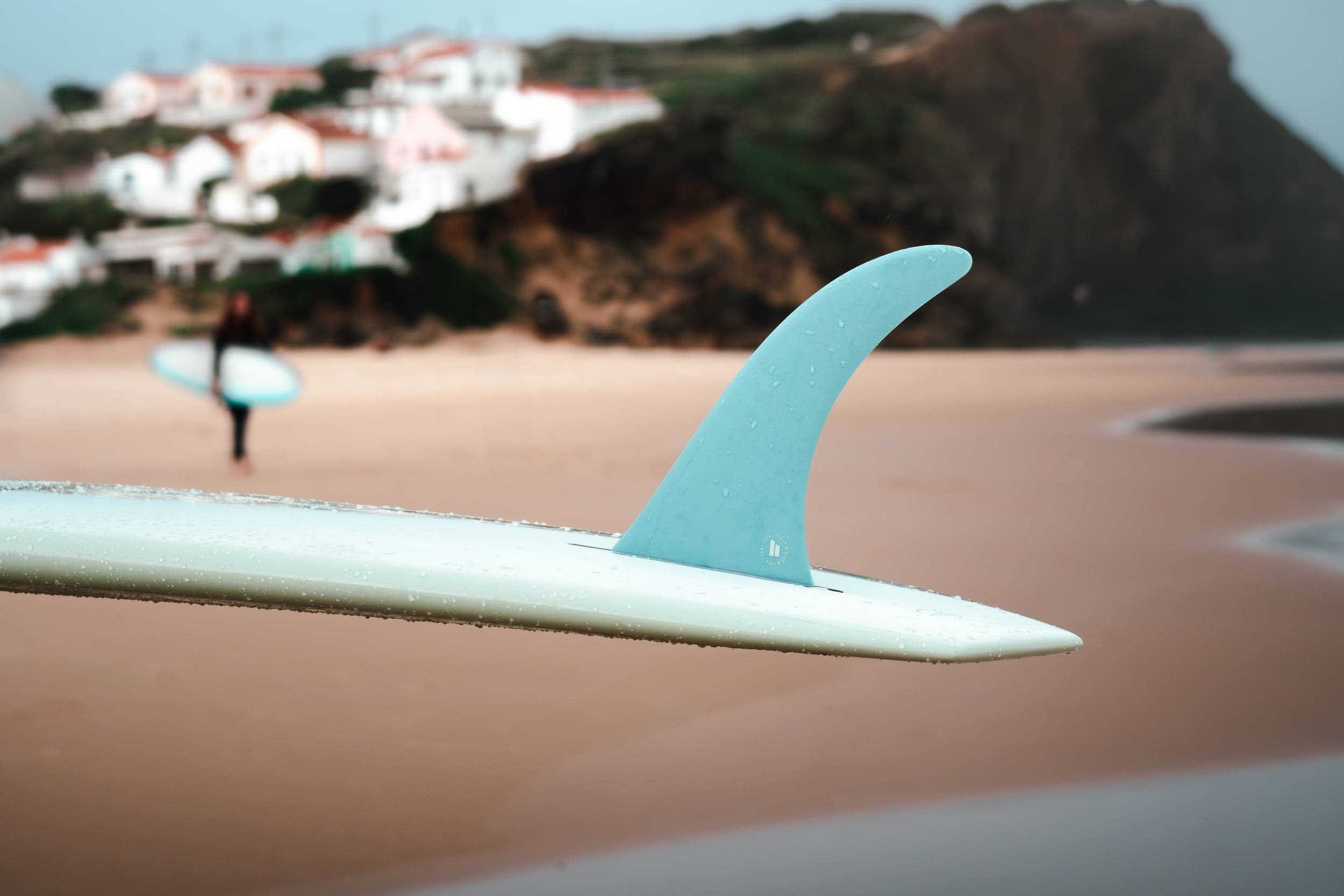 Close-up of a blue surfboard fin, a surfer and Algarve cliffs blurred behind