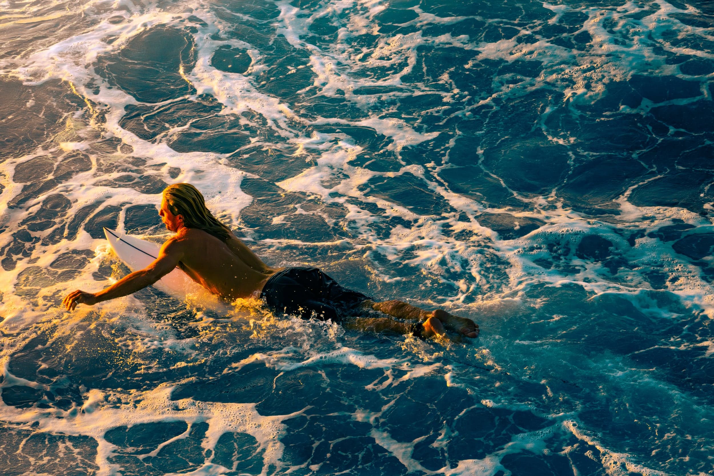 Long-haired surfer paddling across golden foam-streaked water at sunset