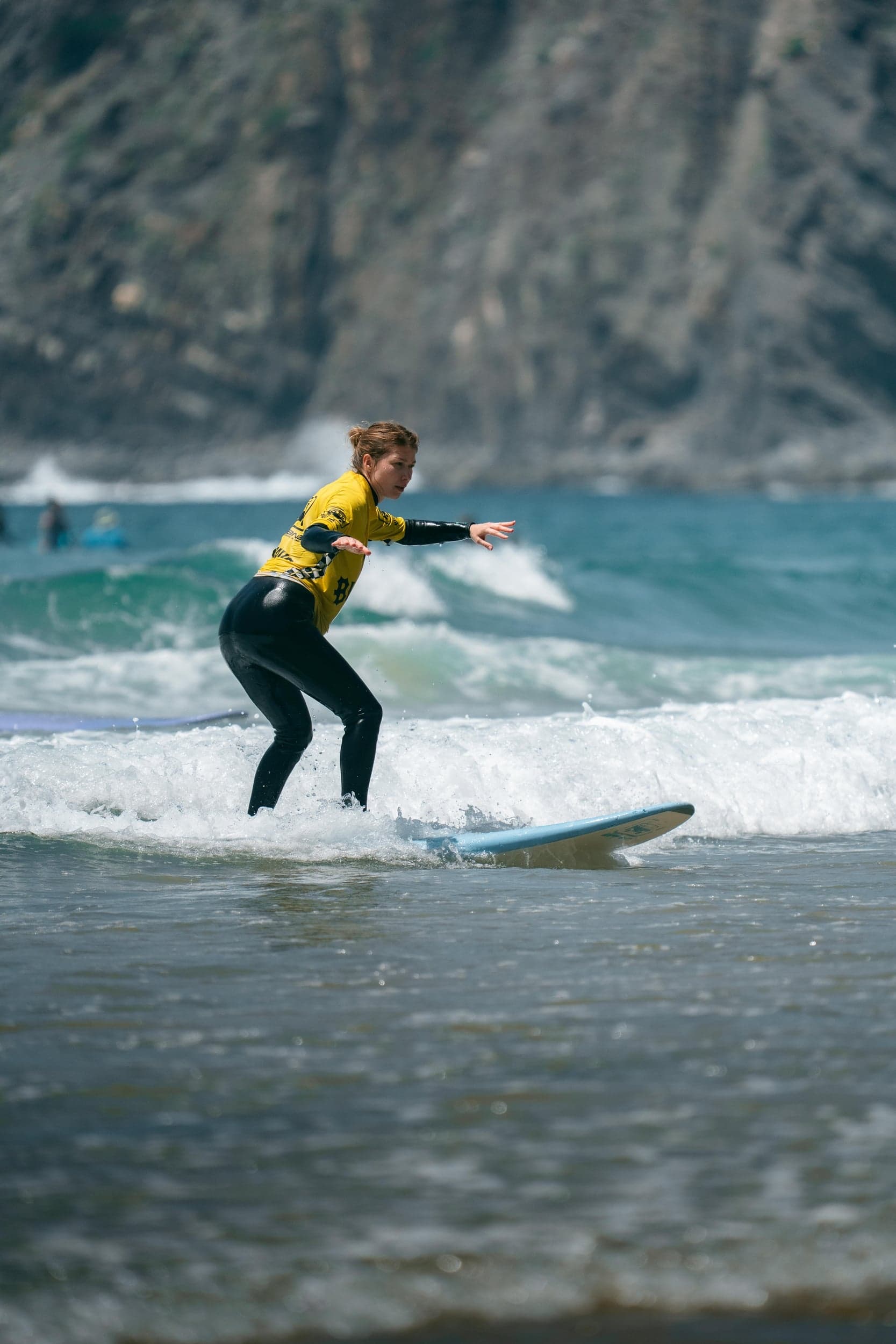 Woman in a yellow rash vest riding a foamboard, rugged cliff rising behind