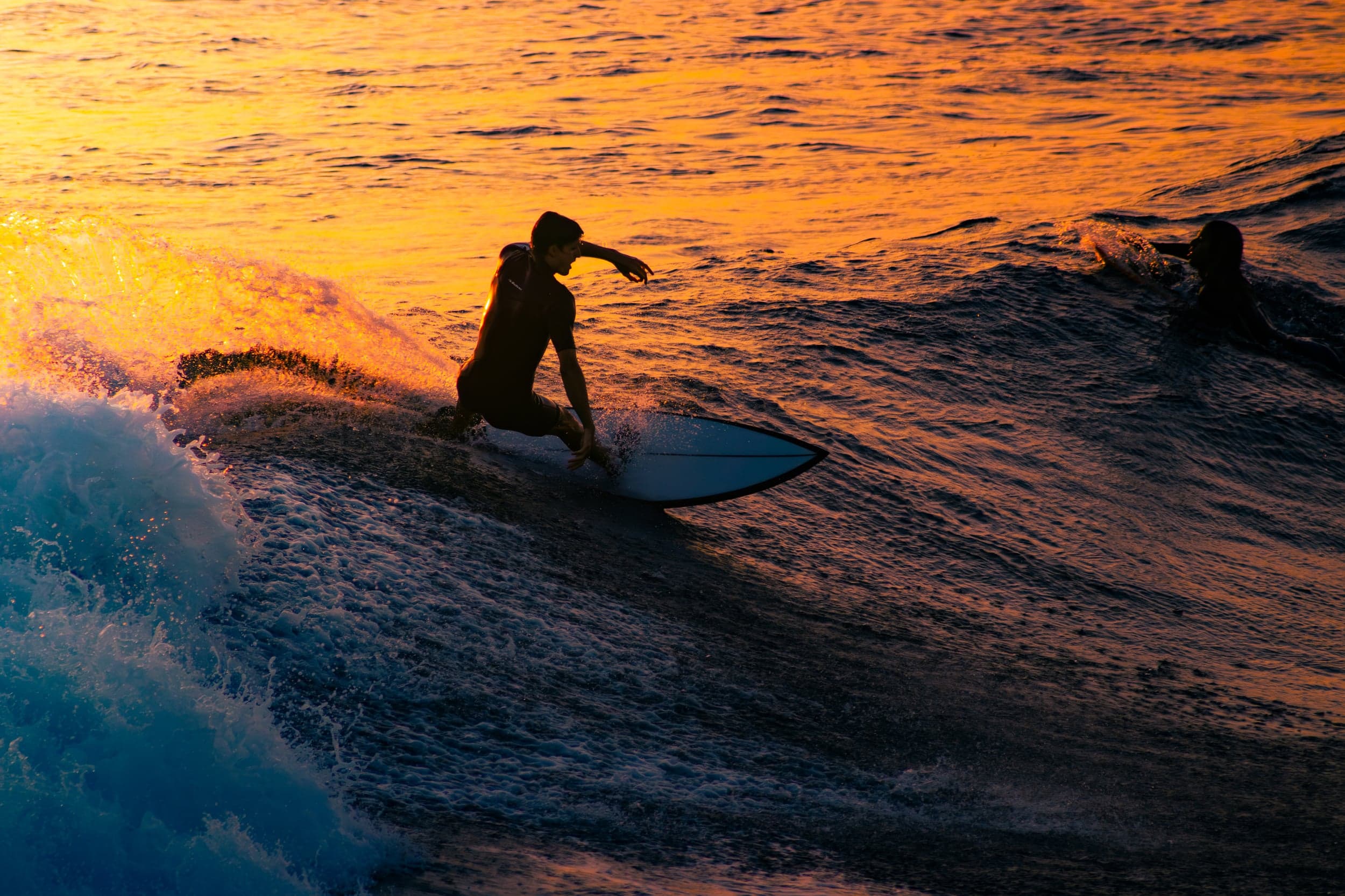 Surfer carving a wave silhouetted against a fiery orange sunset, another surfer paddling behind