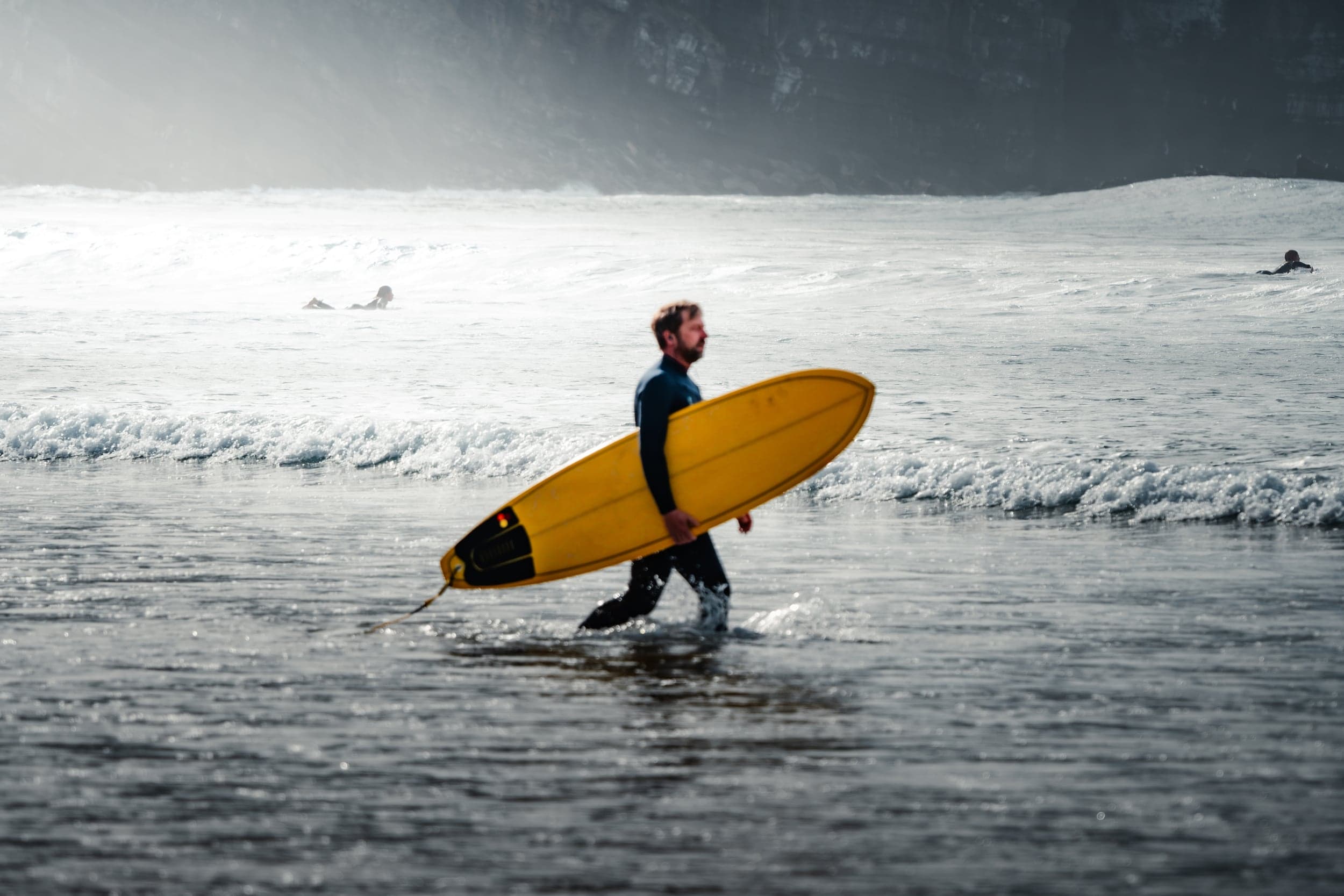 Surfer wading through shallows with a yellow board, misty cliff behind