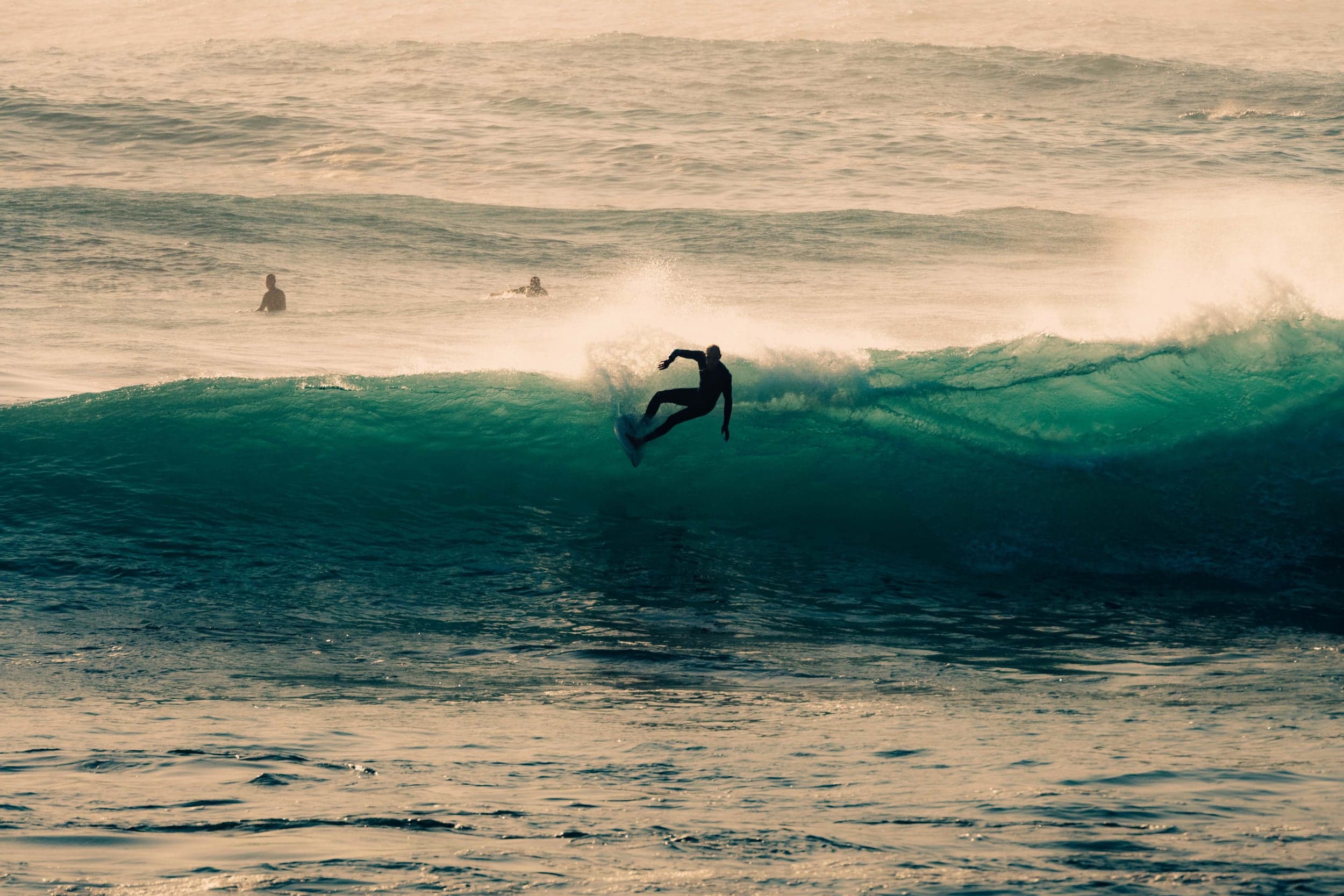 Surfer dropping down a green wave face at sunset, others waiting in the lineup