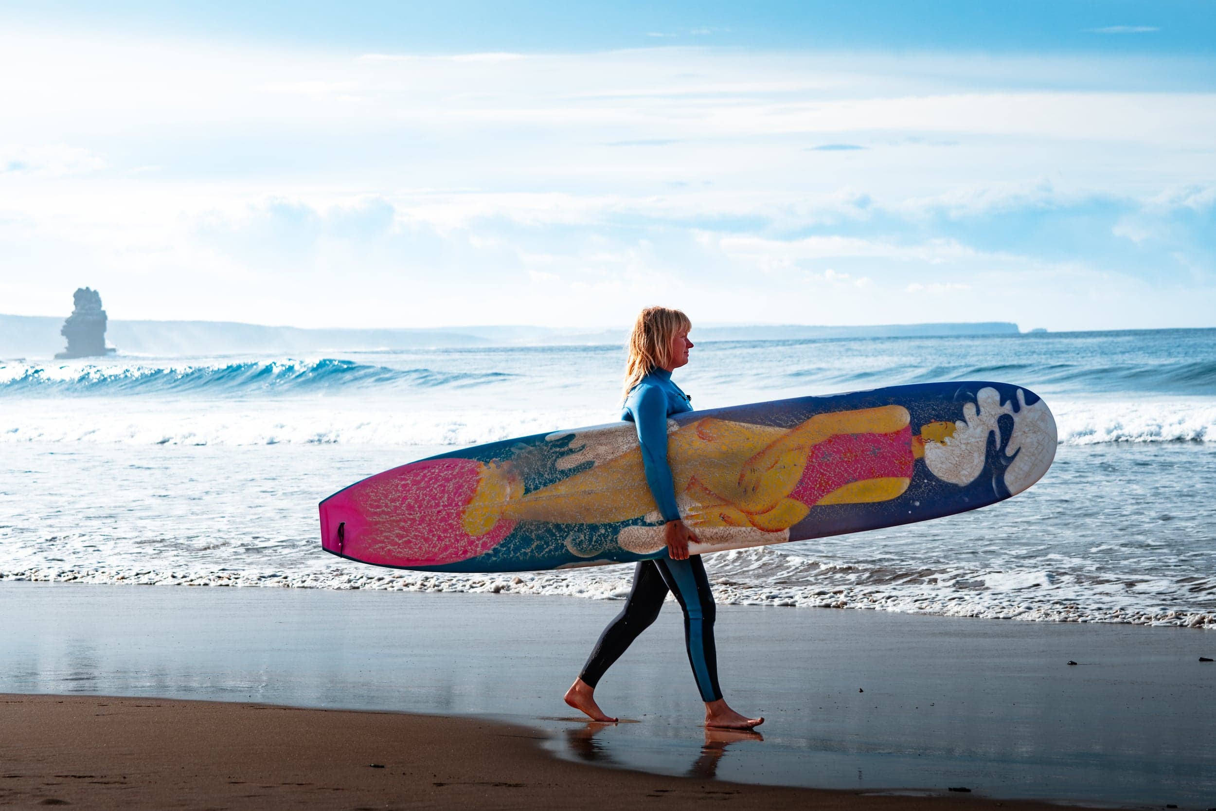 Woman walking the beach with a painted longboard, sea stack on the horizon