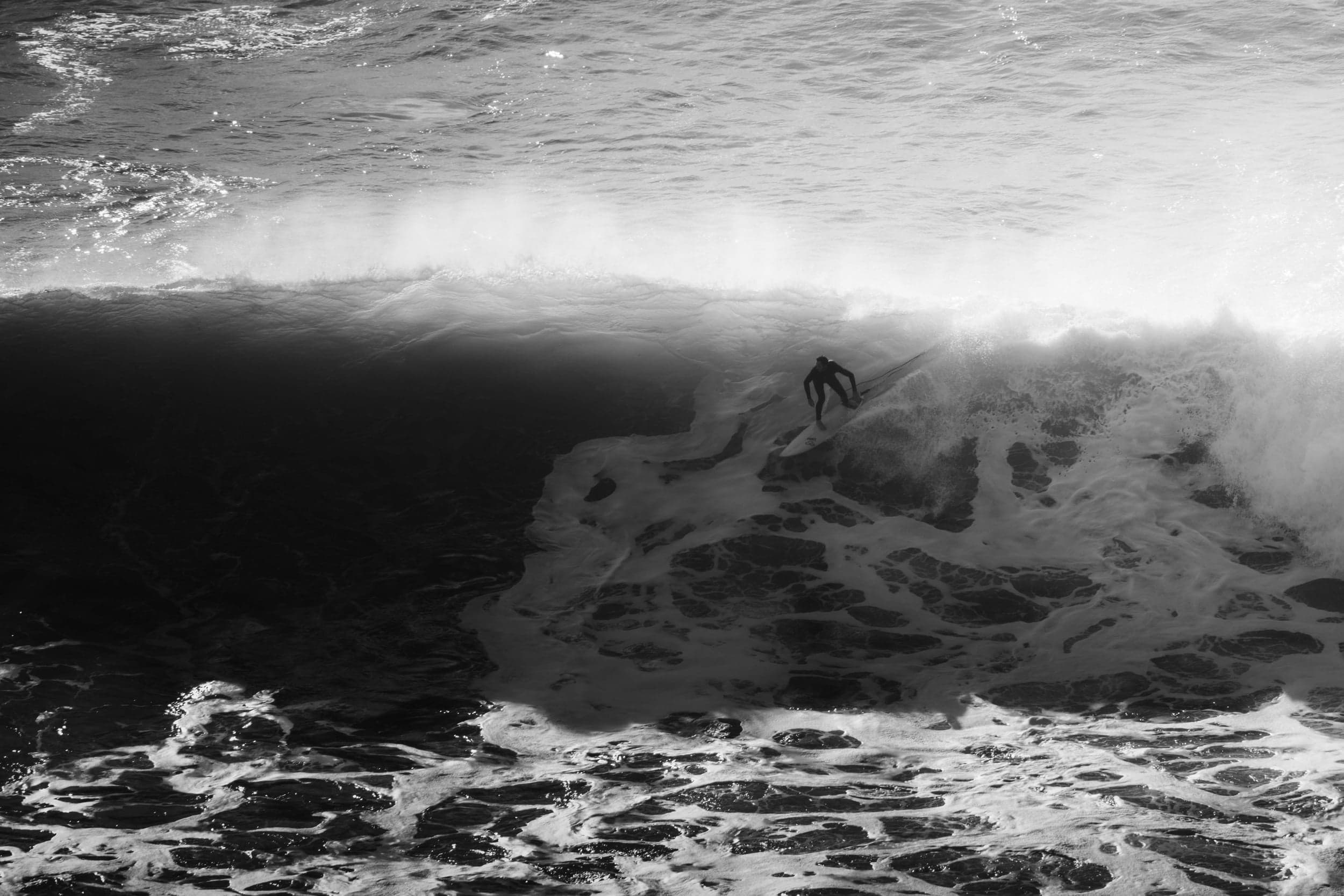 Black-and-white shot of a lone surfer dropping into a dark towering wave, foam swirling below